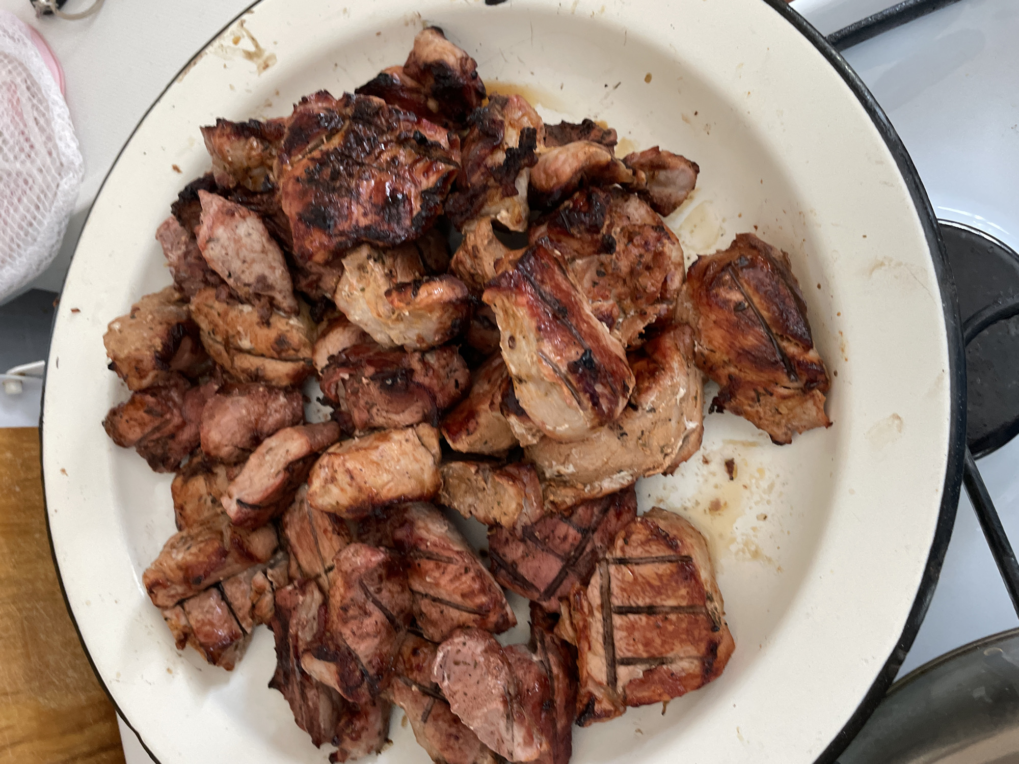 A top-down shot shows a white enamel plate filled with grilled meat pieces, seemingly on a stovetop, possibly in Zapadnaya, Russia. The meat pieces vary in size and shape, displaying grill marks. The plate is old with a black rim and some food stains. The background includes sections of the stove and a wooden surface. No people are visible.