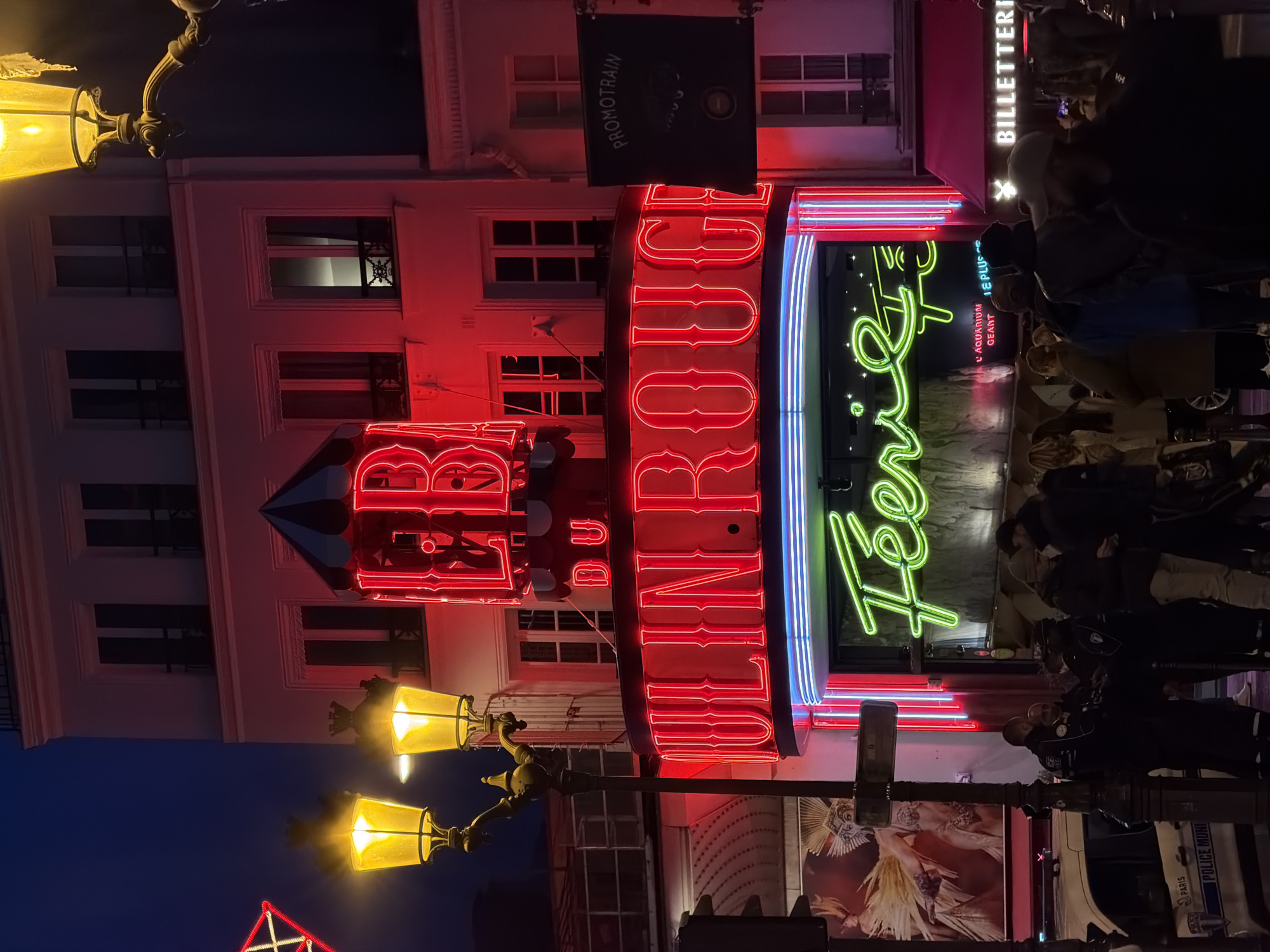 The image captures the exterior of the Moulin Rouge in Paris, France, at night. The building is illuminated by its iconic red neon sign reading "Moulin Rouge" vertically. Below the sign, there is a green neon sign that reads "Feerie," also in vertical format. A black banner with white text, possibly advertising "Promotrain," is hanging on the building's upper levels. A traditional Parisian streetlight with two yellow lamps is visible in the foreground. A crowd of people is gathered near the entrance. A police car is parked along the sidewalk, in front of the crowd.