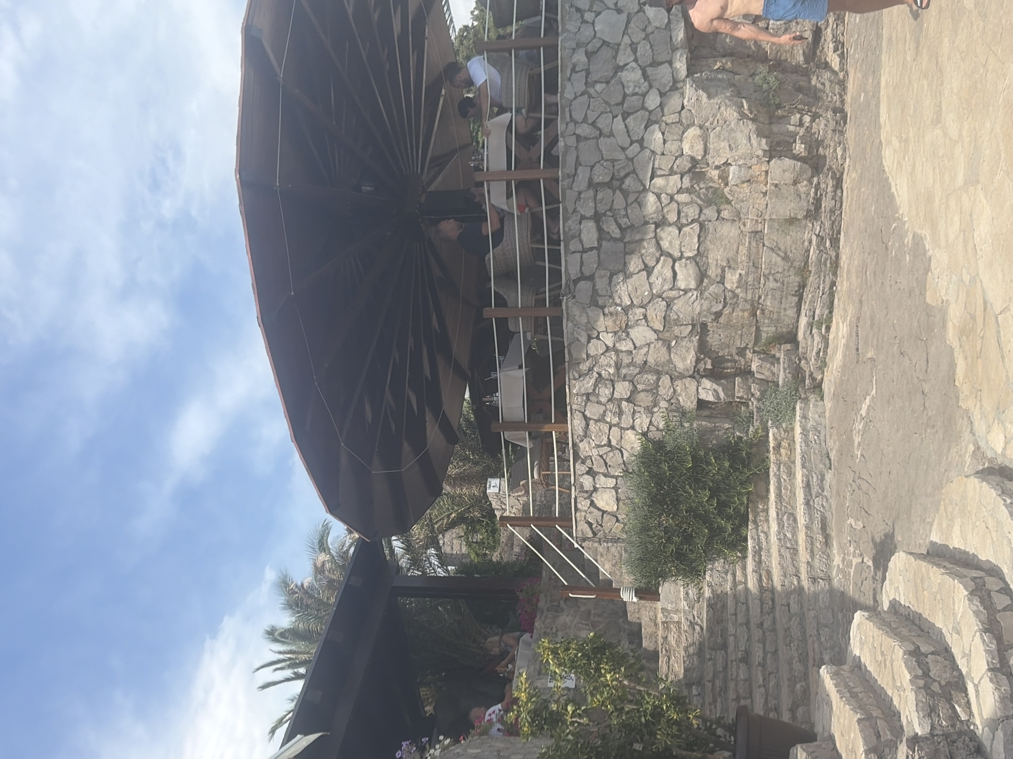 The image depicts an outdoor restaurant situated on a rocky hillside overlooking a body of water, likely the sea or a large bay, given the presence of palm trees and the general ambiance. The restaurant features a large, dark umbrella providing shade over dining tables. Several people are seated at tables, some engaged in conversation, while others appear to be eating. The architecture is characterized by natural stone construction, with a set of stone steps leading up to the restaurant area. Lush greenery, including bushes and flowering plants, is visible around the dining area and along the steps. The sky is blue with scattered clouds, suggesting a bright, sunny day. The presence of a "TOILET" sign indicates public facilities are available. In the foreground, a man in blue swimming shorts is walking away from the camera. The overall scene suggests a relaxed, upscale dining experience in a scenic coastal location within Prijevor, Montenegro.
