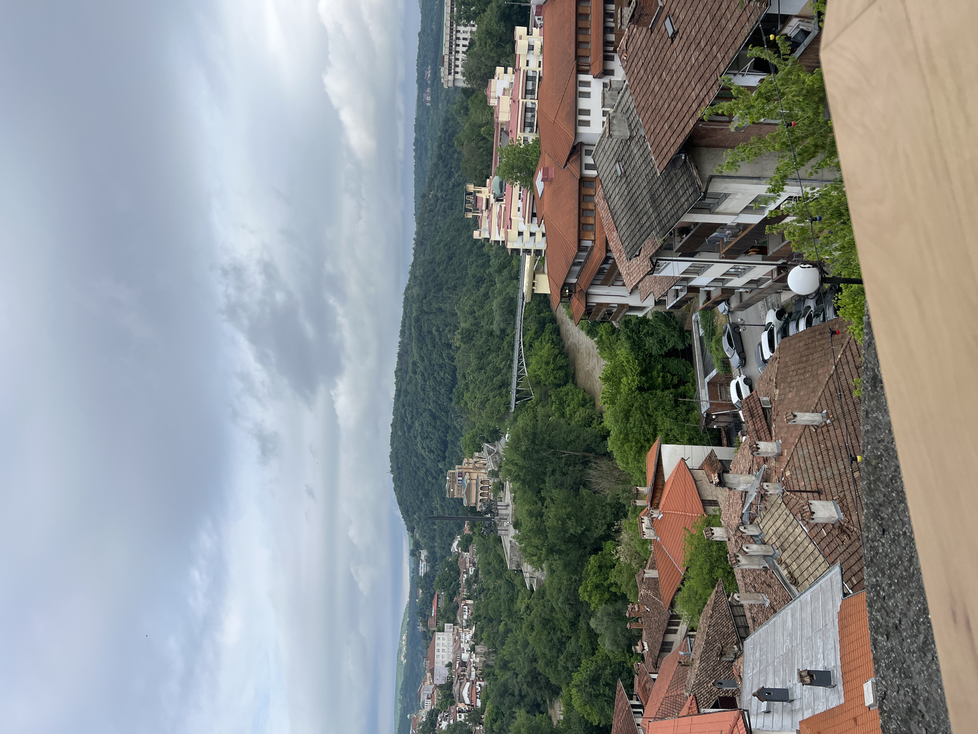 The image shows an elevated view of Veliko Tarnovo, Bulgaria, with a cloudy sky. The foreground displays rooftops with red and grey tiles. Below are tree-covered hills and valley, interspersed with buildings and structures. A bridge spans the valley. Cars are parked along the roads.