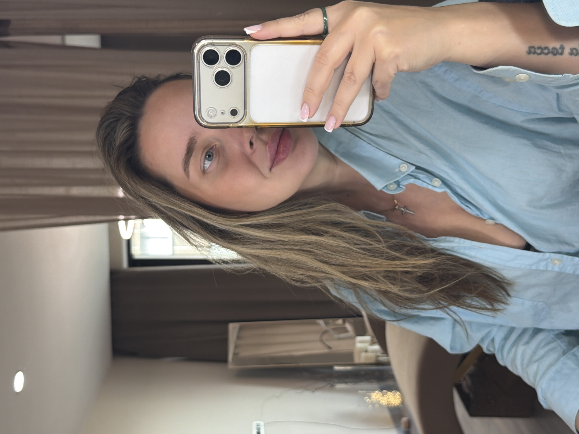 A woman is holding a smartphone with a gold case in front of her face, presumably taking a selfie. She has light skin, blue eyes, and long blonde hair. She is wearing a light blue button-down shirt and a necklace with a cross pendant. Her nails are manicured with a white tip design. She has a ring on one of her fingers. A tattoo is visible on her left arm.
The background appears to be an interior space, possibly a room in an apartment or house. There's a window, a curtain, and what seems to be furniture. Considering the window's appearance and the interior's aesthetic, the image could potentially be taken in a residence or similar setting, but this information is not enough to place the scene in Moscow or elsewhere. Further information is required to make such determination.