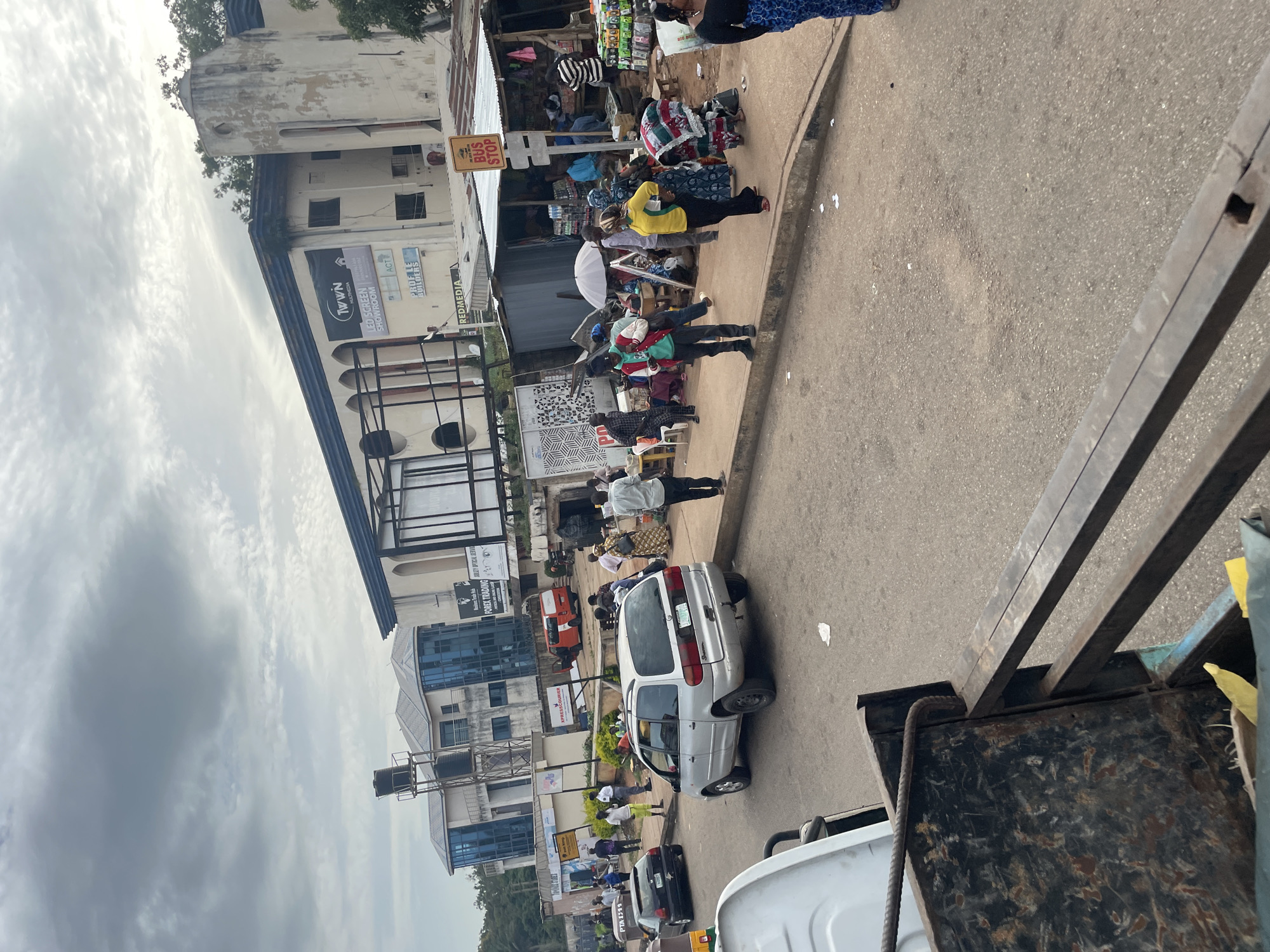 This is a medium-angle, eye-level, outdoor, daytime, full shot of a busy street scene in Jos, Nigeria. In the foreground, the metallic edge of a vehicle's cargo bed is visible, showing signs of wear and tear. The street is paved with asphalt, and a red and white bus is parked in the background. A silver minivan is in the middle ground, facing away from the camera. A black sedan is parked to the left of the minivan.
The sidewalk is lined with various stalls and shops. A white umbrella is set up in front of one stall. People are walking and standing on the sidewalk, some appear to be customers or vendors. A "BUS STOP" sign is prominently displayed. The background features a multi-story building with modern blue windows, and older, weathered structures with archways. The sky is overcast with dramatic clouds. The overall impression is of a bustling urban environment.