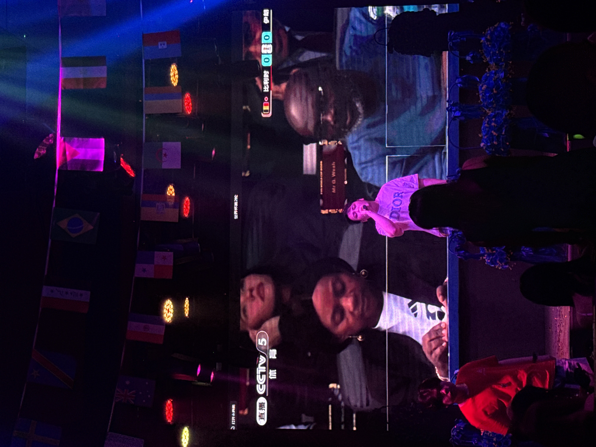In the city of Yiwu, China, a dimly lit indoor event space is captured with a large screen displaying a football match or sporting event. The screen shows several individuals, including a woman in the foreground wearing a white top and a black jacket, and behind her, another woman with dark hair and red lipstick. The screen also features graphics related to the event, including "CCTV 5" and a score that reads "0-0." A third individual, a Black man with glasses and a beard, is visible on the screen. In front of the screen, a young person wearing a pink t-shirt with "DIOR" written on it is singing into a microphone. To the left of the screen, a young person in a red t-shirt is standing. The environment is decorated with numerous small national flags strung across the ceiling and colorful stage lighting. In the foreground, the silhouettes of audience members are visible.