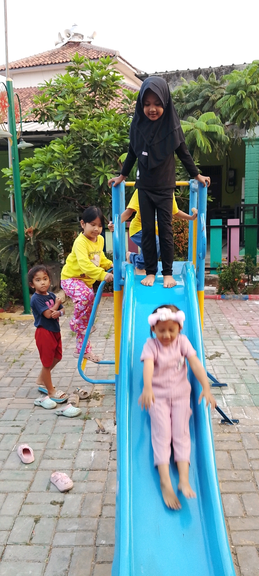 In Satriamekar, Indonesia, a blue and yellow playground slide is the focal point of the scene. A young girl wearing a pink outfit and a pink headband is actively sliding down the bright blue slide, her motion blurred by speed. To the left, another young girl in a yellow long-sleeved shirt and patterned pink pants stands on the middle platform of the slide, holding onto the blue railing. Beside her, at the base of the slide structure, a third young girl in a dark blue t-shirt and red shorts stands with her arms crossed, looking towards the left. A fourth child, partially visible, is on the top platform of the slide, wearing a black hijab and black clothing. Surrounding the playground equipment is a brick-paved area with scattered sandals and flip-flops. Lush green trees and foliage frame the background, with a building featuring a tiled roof and a prominent white structure, possibly a speaker system, visible in the distance.