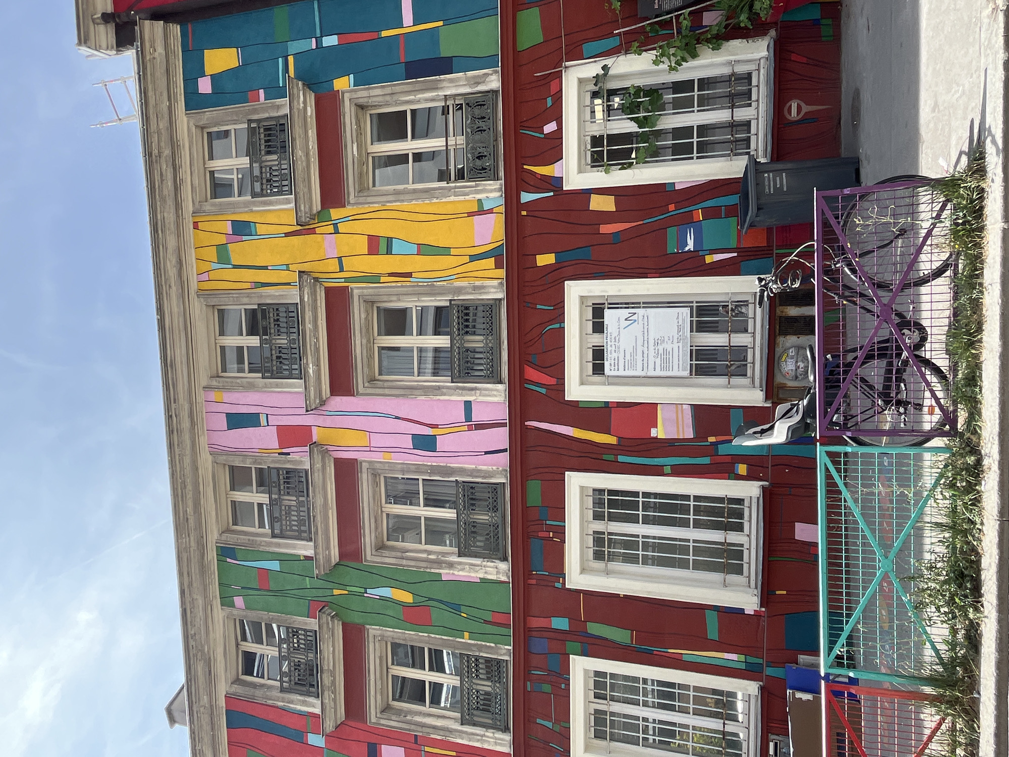 This image shows a colorful building facade on a sunny day in Paris, France. The building is adorned with a vibrant mural featuring abstract, vertical lines of various colors, including red, green, yellow, pink, and blue. The mural extends across multiple stories, encompassing several windows on each level. The windows on the upper floors are smaller and feature black wrought-iron balconies. The windows on the lower floor are larger and have white frames with gridded panes.
In front of the building, a sidewalk runs along a street. A brightly colored metal fence, segmented into red, teal, and purple sections, separates the sidewalk from the building. A bicycle with a purple frame is parked behind the fence. A dark gray trash bin stands near the building, to the right of a window. A white sign with text is posted on the window to the left of the trash bin. Green vines are growing up the side of the building near the right edge of the frame. The sky above is blue with a few wispy clouds. Scaffolding can be seen on the roof of a neighboring building on the right side of the image.