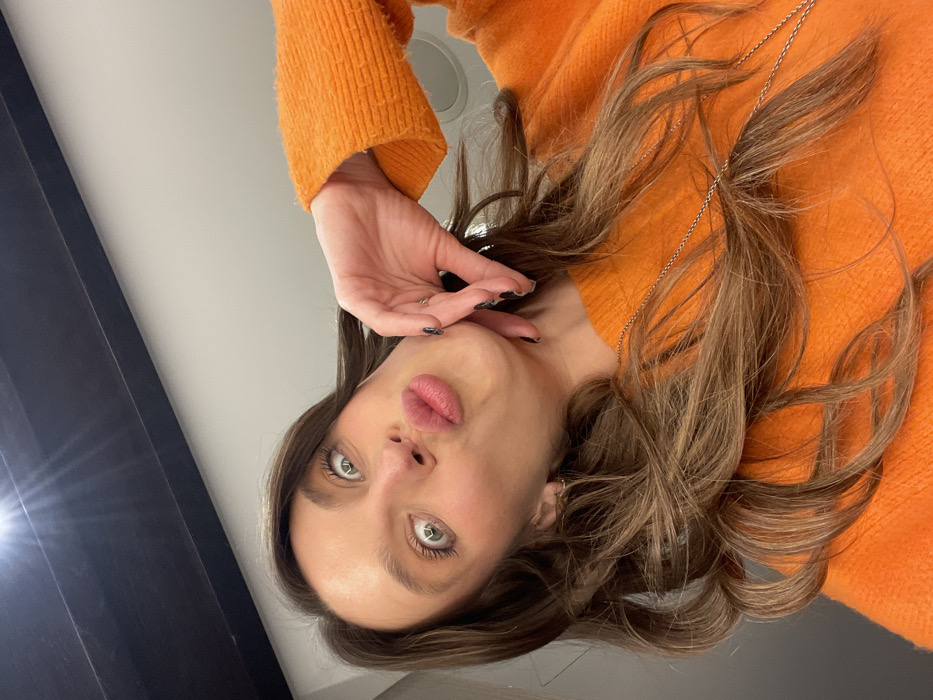 A woman with long brown hair and green eyes poses in a bright orange sweater. Her lips are puckered, and her right hand is near her face. She is wearing a silver necklace. The photo is angled so that the woman's face is at the bottom. The background includes a light gray wall and a dark blue curtain or doorframe. The lighting is bright with some reflections on the dark surface.