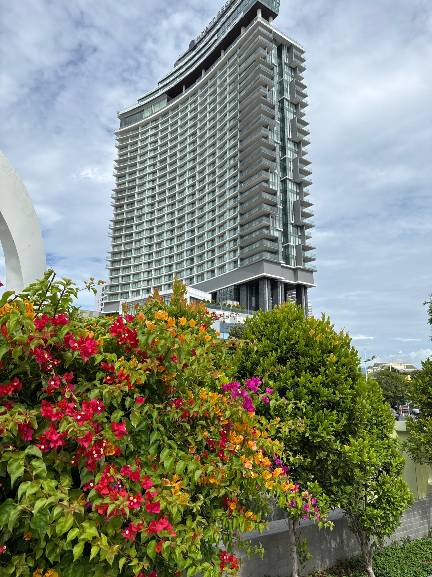 The image shows the Hyatt Regency Nha Trang hotel, a tall, modern building with a curved facade of balconies and windows, situated in the city of Nha Trang, Vietnam. In the foreground, vibrant bougainvillea flowers in shades of red, pink, and yellow bloom against lush green foliage. The sky is overcast with white clouds. A large white curved structure is partially visible on the left side of the image. The scene appears to be outdoors, likely a public space or hotel grounds. There are no people visible in the image.
