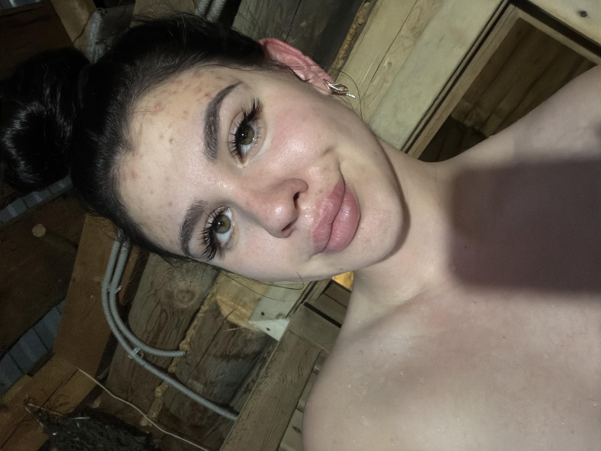 A young woman with dark hair pulled up in a bun is taking a selfie in what appears to be a wooden sauna. She has light brown eyes, full lips, and some acne on her forehead and cheeks. She is wearing a silver earring with a diamond accent in her right ear. Her shoulders and upper chest are visible and appear to be glistening, suggesting she is sweating. The background consists of wooden walls and ceiling, with some electrical conduits visible. The lighting is warm and intimate, casting shadows on the wooden surfaces. The overall impression is that of a personal moment captured in a private, rustic setting. The location is not specified in the image, and therefore its presence in the city of Фоки, Russia, cannot be confirmed.