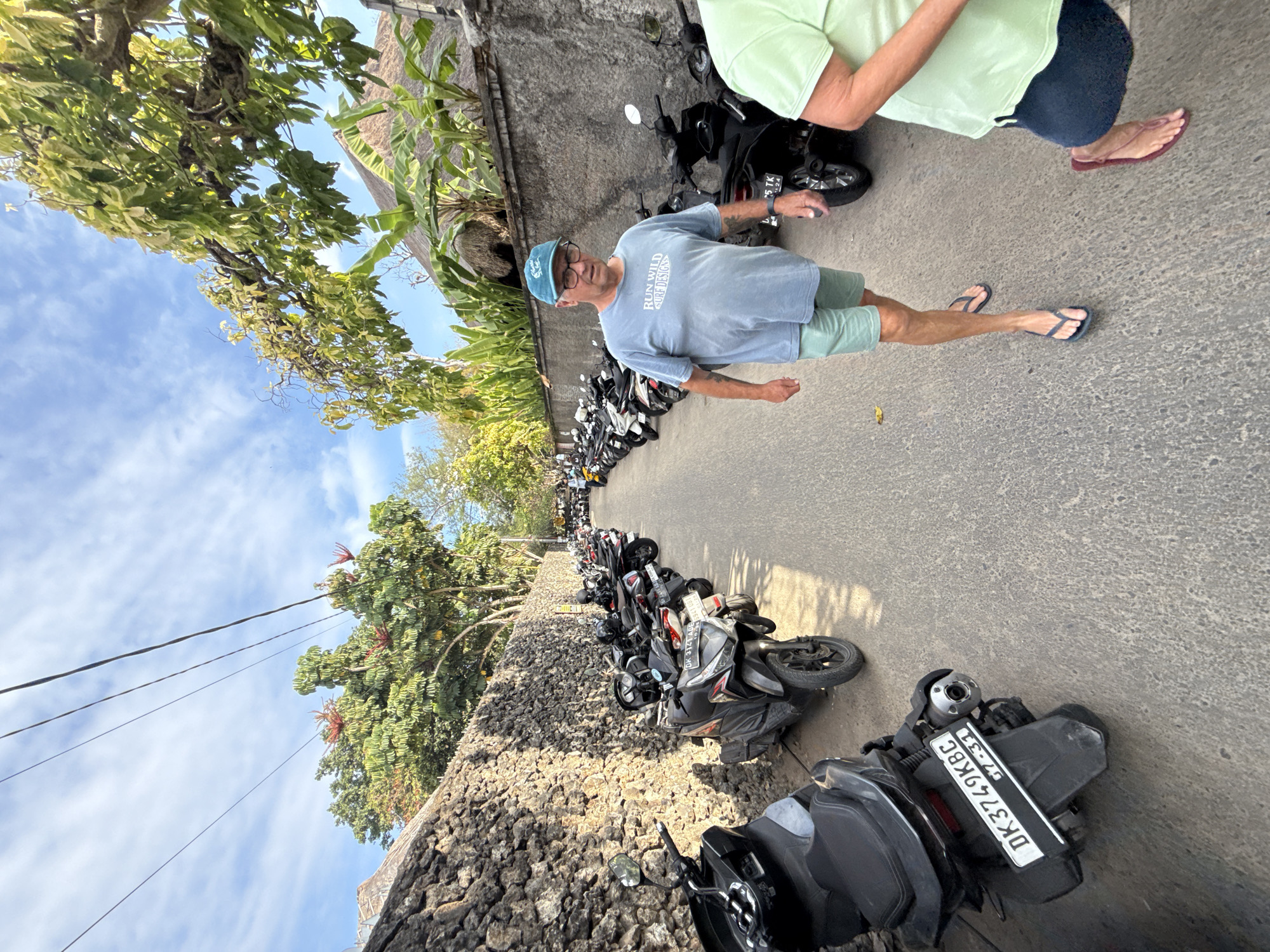 A medium shot captures a street scene in Denpasar, Indonesia, featuring a man in a blue t-shirt and light green shorts walking towards the camera. He wears a blue cap and glasses. A parked line of scooters runs along the left side of the narrow street, bordered by a rough stone wall. Another scooter is parked on the right. Behind the man, other scooters and people are visible further down the street. Banana trees and foliage sprout from the top of the wall on the right, and trees with red blossoms and green leaves are visible on the left. The sky above is blue with scattered white clouds. The man's t-shirt has text that reads "RUN WILD SURF DESIGN". The license plate on the closest scooter is partially visible, showing "DK3749KBC 07-31".