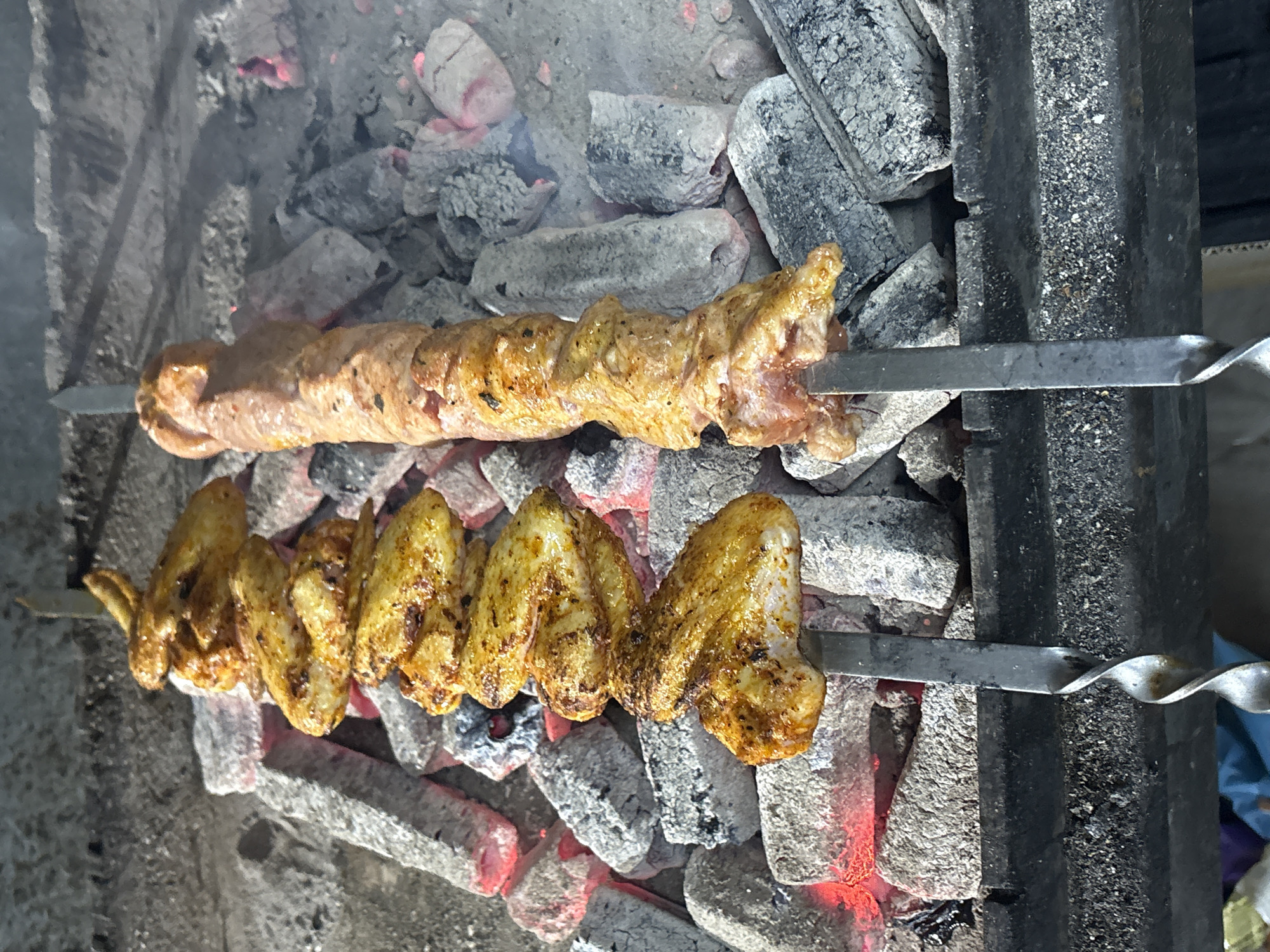 The image shows two skewers of food grilling over hot coals. The top skewer holds a sausage-like meat product. The bottom skewer contains several chicken wings. The coals are arranged beneath the skewers, and some embers are visible. The location is unknown.