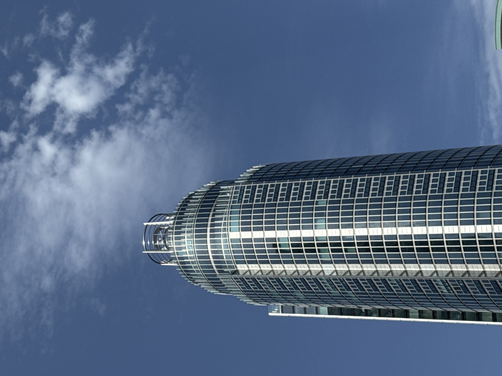 A low-angle, close-up shot focuses on a contemporary glass skyscraper set against a clear blue sky with sparse white clouds. The building features a curved, cylindrical facade composed of numerous rectangular windows framed in white. The top of the skyscraper has a distinctive circular railing. To the left of the main building, a smaller, slimmer building with a similar glass facade is partially visible. The image does not depict any people or specific landmarks that would indicate its location in London, United Kingdom.