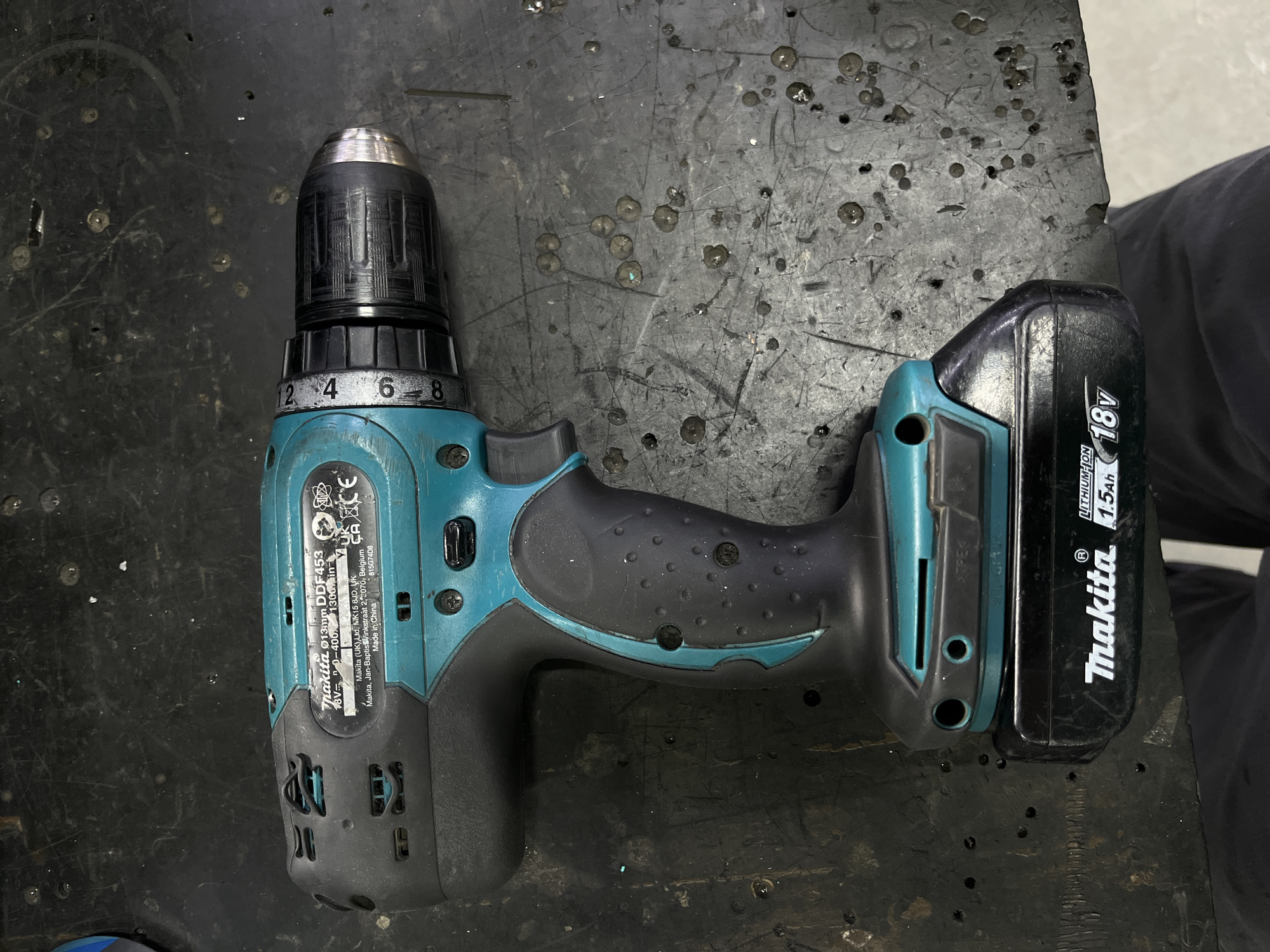 A Makita DDf453 cordless drill with an 18V 1.5Ah battery is displayed on a dark, worn, and textured surface. The drill is teal and black with a silver chuck. Visible markings on the drill indicate it is made in China and its model number and specifications. The surface appears to be a workbench or table with various dark spots and marks, possibly from use or spills. The background is dark and out of focus, obscuring any specific details of the surroundings. No people or specific actions are depicted. The image is taken from a top-down perspective. There is no identifiable information regarding the location in Kyiv, Ukraine.