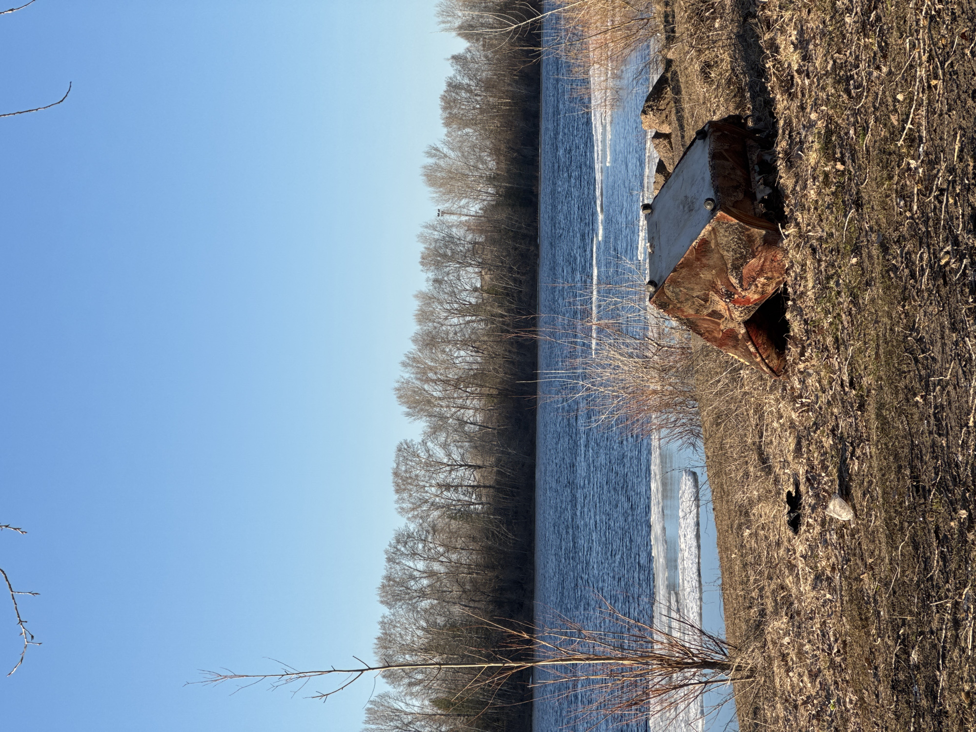 The image depicts a riverbank scene in an outdoor setting. The river's water is visible, with trees lining the opposite bank. A large, rusty metal object, possibly a container or piece of equipment, sits on the riverbank among dried vegetation and debris. The sky is clear blue. The location is plausibly near Chaikovsky, Russia, given the typical Russian landscape of rivers and forests. No people are visible. The focus is on the discarded metal object in the natural environment.