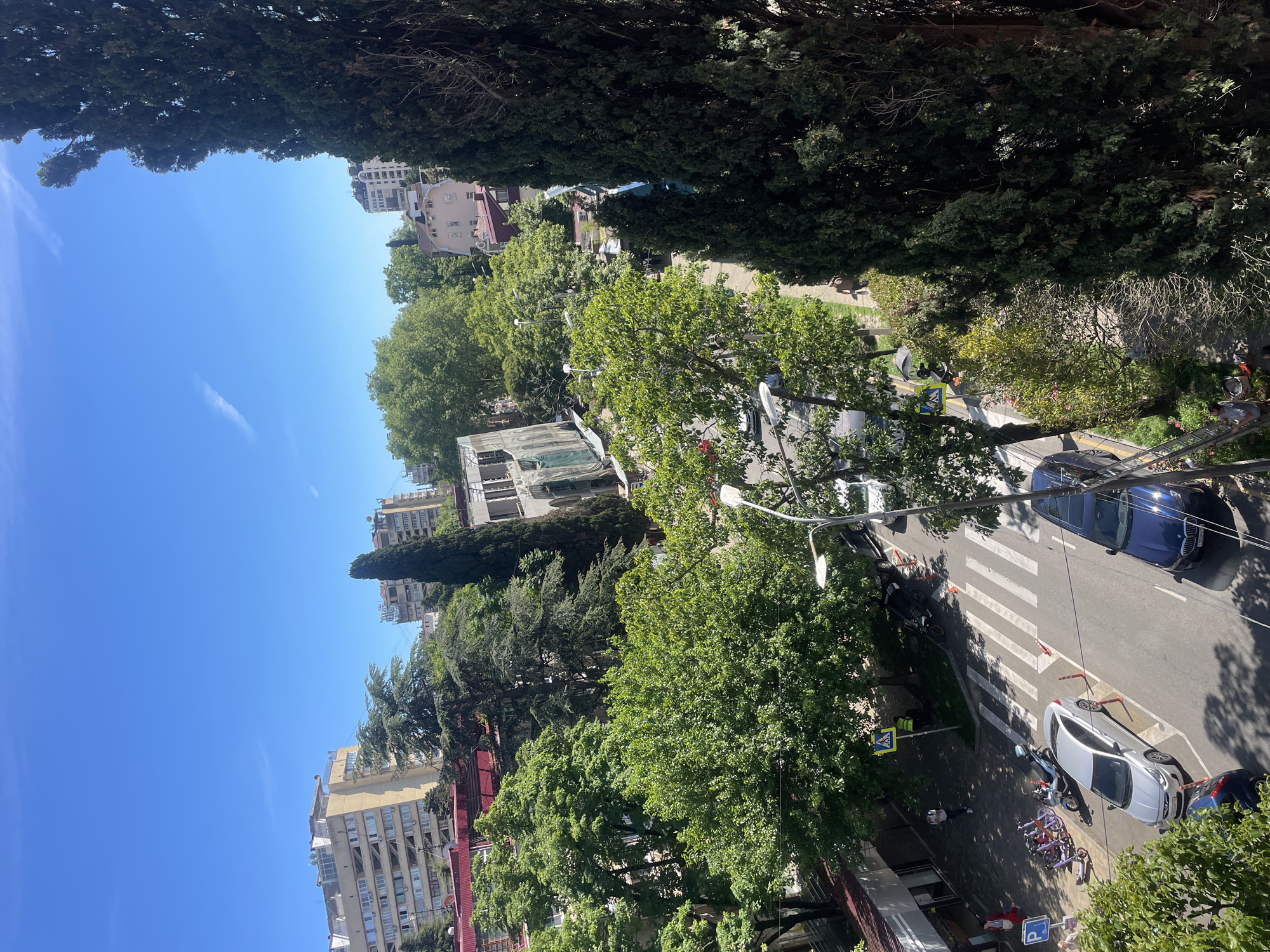 This image provides an elevated, overhead view of a bustling urban street scene in Sochi, Russia, under clear blue skies. The perspective looks down onto a vibrant area characterized by a mix of buildings, a street, and abundant green trees.
The sky, filling the upper left portion of the image, is a bright, clear blue, with a single faint contrail visible. Tall, dark green trees frame the scene, suggesting a verdant environment. Along the street, numerous trees with full, lush green canopies line the sidewalks and roadways, providing significant shade.
On the left side of the street, multi-story buildings, varying in architectural style and height, are visible. Some feature balconies and rooftop satellite dishes. The street itself, asphalted, contains a clearly marked pedestrian crosswalk. Several vehicles are present: a dark blue SUV is seen towards the upper right, while a silver sedan is parked on the right sidewalk. A person is visible on a ladder next to the silver car, suggesting some activity or maintenance. Other parked vehicles include a motorbike/scooter, bicycles, and purple electric scooters. Orange traffic cones are placed near the silver car and the crosswalk area.
Several people are scattered along the sidewalks; one person in a yellow vest is positioned near the pedestrian crossing sign, which is yellow with a black walking figure. The scene conveys a sense of routine daily life in a sunny, green urban setting. A blue parking sign displaying a white "P" is visible near the parked vehicles. The time of day appears to be midday or early afternoon, indicated by the bright sunlight and distinct shadows.