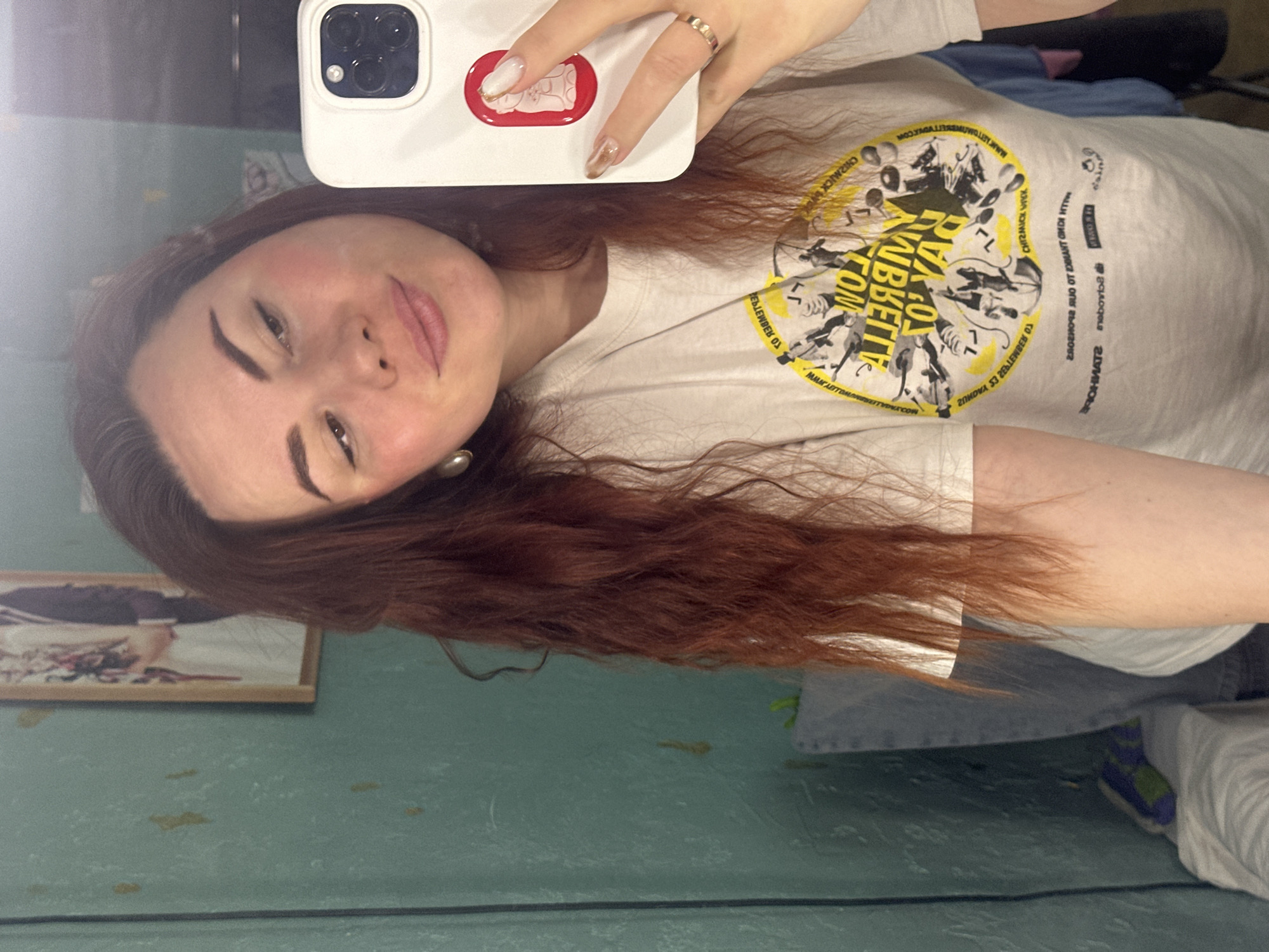 A woman is taking a selfie, holding a white iPhone with a red sticker on the back. She has auburn hair, fair skin, and dark eyebrows. She's wearing a white t-shirt with a yellow and black graphic design. A picture frame hangs on a green wall, likely in Черёмушки, Russia. The background shows a glimpse of casual clothing. The text on the t-shirt includes the words "TO," "COM," "CHIZMICK," and "MAF LETTOALETYPECON." Other text is reflected, reversed and illegible.