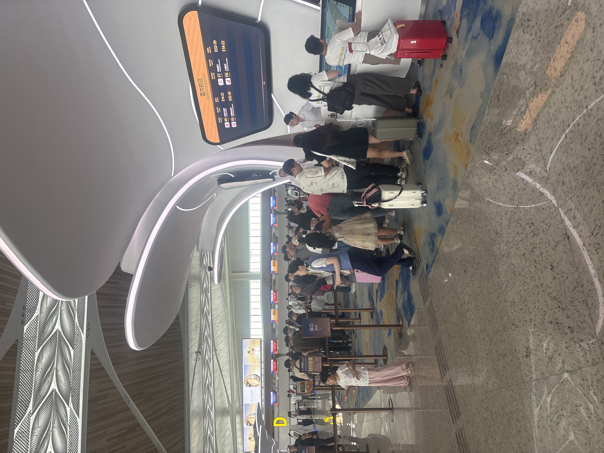 The image depicts a bustling airport terminal in Baiyun District, China, with several queues of passengers. In the foreground, a young girl in a white t-shirt and pink pants stands patiently, facing left, with a stroller behind her. To her right, a woman in a blue uniform, likely an airport employee, walks with a pink suitcase. Further back, more people are lined up at counters, some interacting with staff. Overhead, the ceiling features a modern design with curved white structures and illuminated elements, complemented by wooden accents and a perforated metal pattern. A large digital display board shows "DOMESTIC FLIGHTS" and flight information. The floor is a reflective polished surface with a colorful abstract design in shades of blue and yellow. Several pieces of luggage, including suitcases and bags, are visible around the passengers. The scene suggests a busy departure or check-in area within the airport.