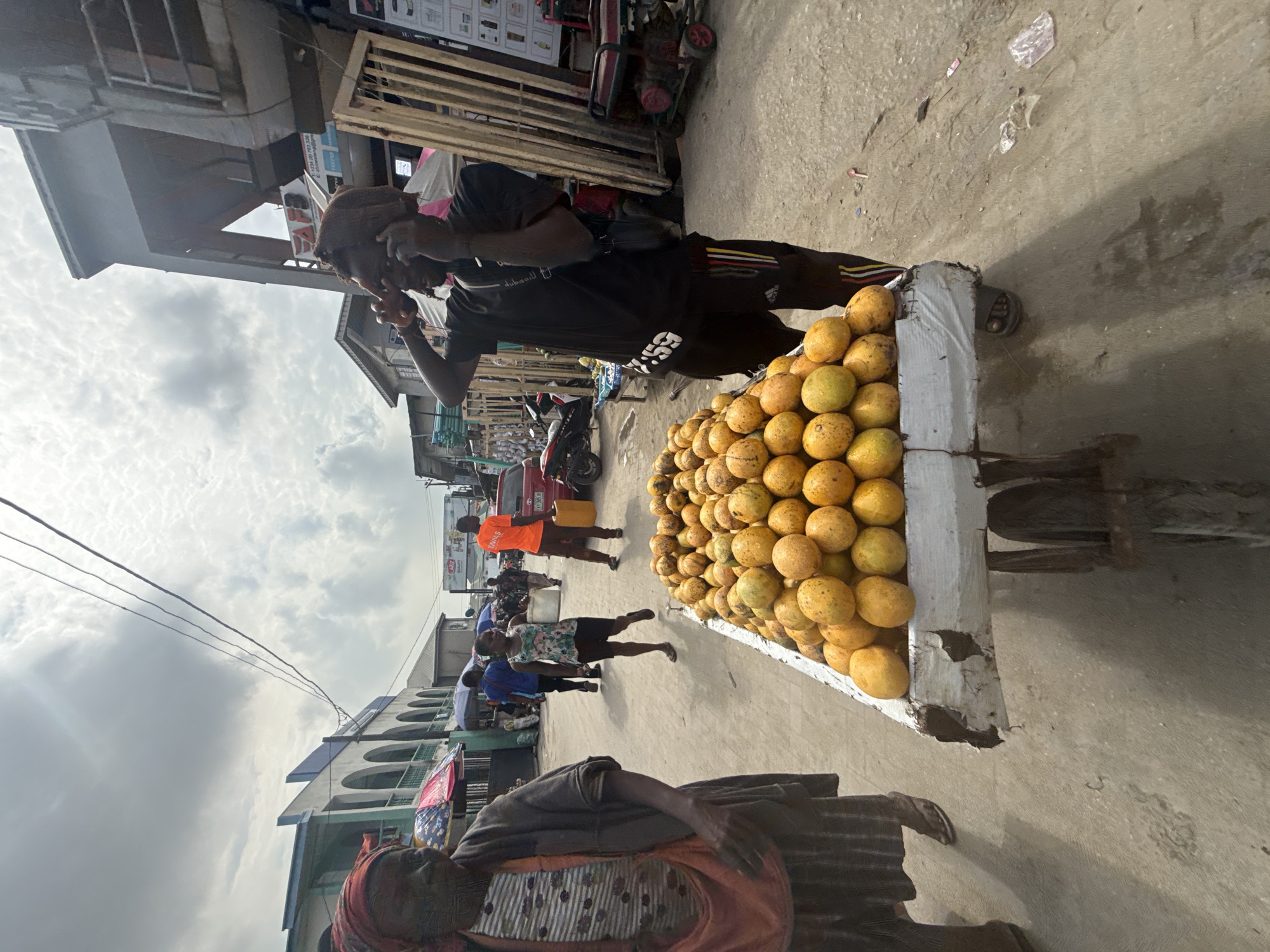 This image captures a bustling street scene in Lagos, Nigeria, on an overcast day. The primary subject is a man standing beside a large, rustic wooden cart overflowing with a heap of round, yellowish fruits, likely oranges or a similar citrus variety. He is wearing a black t-shirt with "55" printed on it and black shorts with red and yellow stripes. The man appears to be speaking on a phone or to someone, with his hand raised towards his ear.
In the immediate foreground, partially cut off, an older woman dressed in patterned traditional attire is visible. Further down the street, several other people can be seen, moving or standing. One person in an orange t-shirt and another in a patterned top are distinguishable among the crowd. Buildings with a typical urban West African architectural style, some featuring arched windows, line both sides of the street. Overhead, power lines crisscross against a cloudy, bright sky. A red motorcycle is parked in the mid-ground. The street itself appears to be a mix of paved and unpaved surfaces, with some litter scattered on the ground near the fruit cart. The scene depicts everyday activity in a lively urban market or commercial area during daytime. No strong emotions are discernible; people appear to be engaged in their daily routines.