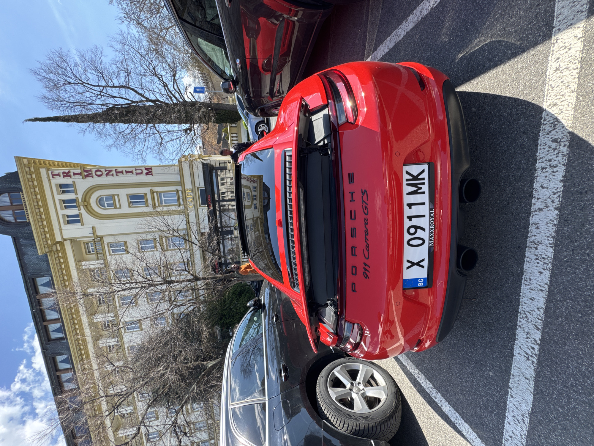 A red Porsche 911 Carrera GTS is parked in a parking spot in front of the Trimontium Hotel Plovdiv, Bulgaria. The car's trunk is open, revealing the engine compartment. The Bulgarian license plate reads "X 0911 MK." Other cars are parked nearby, a black SUV to the left, and another dark-colored vehicle above the Porsche. A person is visible near the open trunk of the Porsche. The surrounding environment includes bare trees and a building with visible architectural details. The time of day appears to be daytime, under bright sunlight.