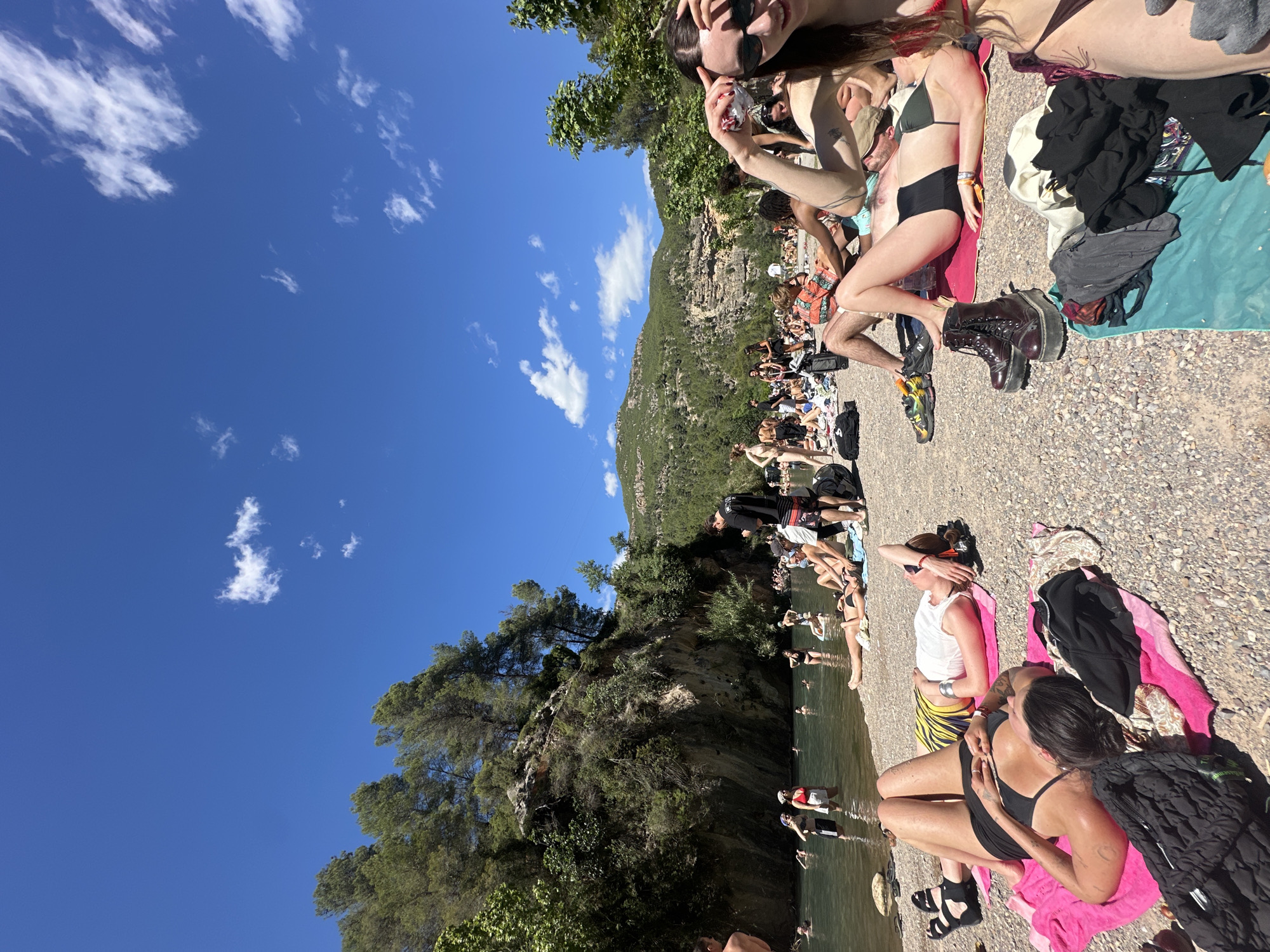 The image shows a crowded riverbank scene in Montanejos, Spain. The river is lined with people sunbathing and swimming. Most people wear swimwear or casual clothing. The river is green, and there are trees and a rocky hill on the other side. Some people are lying on towels on the riverbank. The sky is blue with scattered clouds. The riverbank is made of gravel. Some people are wading or swimming in the river.