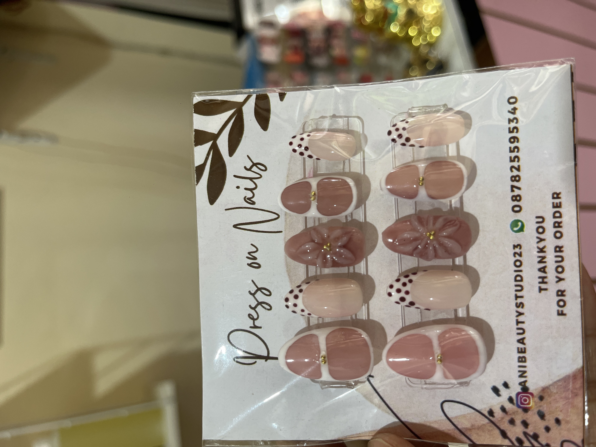 The image captures a package of "Press on Nails" held up by a person's hand in an indoor retail setting, likely a beauty or accessory store. The package contains ten artificial nails, displayed on a white card and sealed in clear plastic. The nails feature designs in nude, light pink, and dusky rose tones. Notable details include nails with dark brown polka dots on a light tip, others with small gold studs, and some with raised, three-dimensional floral patterns.
Prominent text on the left side of the packaging reads "Press on Nails." On the right, contact information is visible, including an Instagram handle "@ANIBEAUTYSTUDIO23" and a WhatsApp phone number "087825595340." Below these details, the phrase "THANKYOU FOR YOUR ORDER" is printed.
The background is out of focus, revealing warm-toned surfaces, possibly shelving or walls, and blurred golden lights, suggesting decorative elements or other merchandise. The scene conveys a moment of product display or examination within a retail environment, consistent with a beauty product shop in North Kuta, Indonesia.