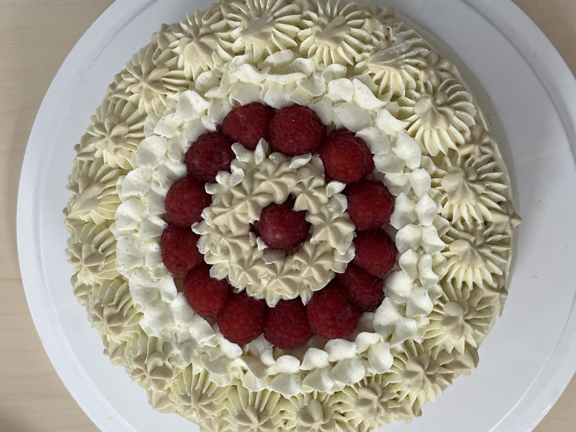 A close-up, top-down view captures a round cake presented on a white cake stand. The cake is adorned with decorative frosting and fresh raspberries. The frosting is applied in concentric circles, featuring a combination of star-shaped piped designs in a light beige color, interspersed with dollops of white frosting. Fresh, vibrant red raspberries are strategically placed within the frosting patterns. The background appears to be a light-colored, possibly wooden surface. The image does not contain any elements that would suggest a specific location in Plovdiv, Bulgaria.