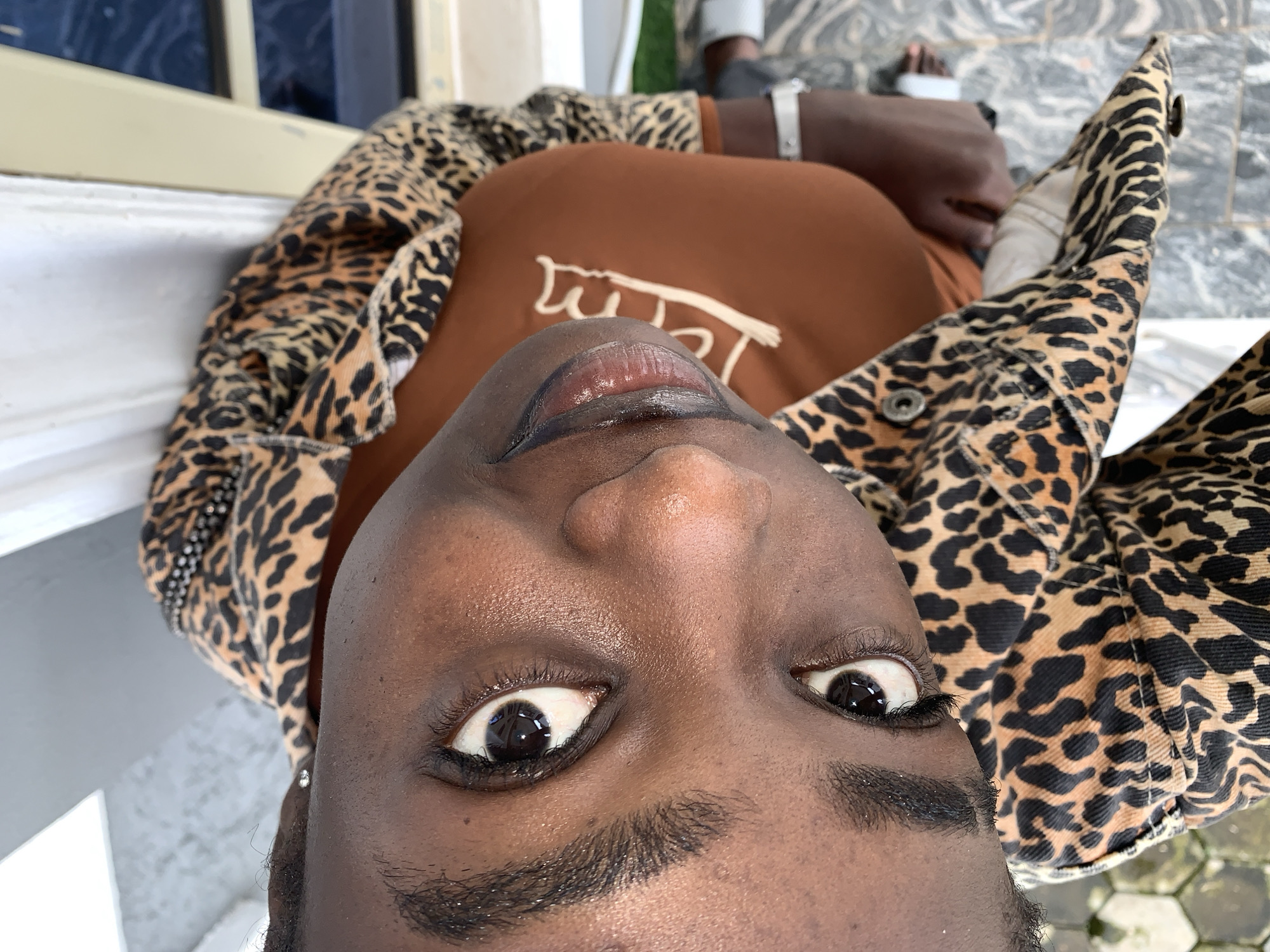 A close-up, portrait-style, sideways shot shows a person's face and upper body. The person has dark skin and is wearing a leopard-print jacket over a brown shirt with a white graphic. The background is blurred, but appears to be an outdoor setting with a tiled floor and a building. The image is cropped and rotated, making it difficult to determine specific details about the environment or location within Gura Top, Nigeria. The lighting is natural, suggesting it was taken during the day. The person's eyes are looking directly at the camera, and their lips are slightly parted.