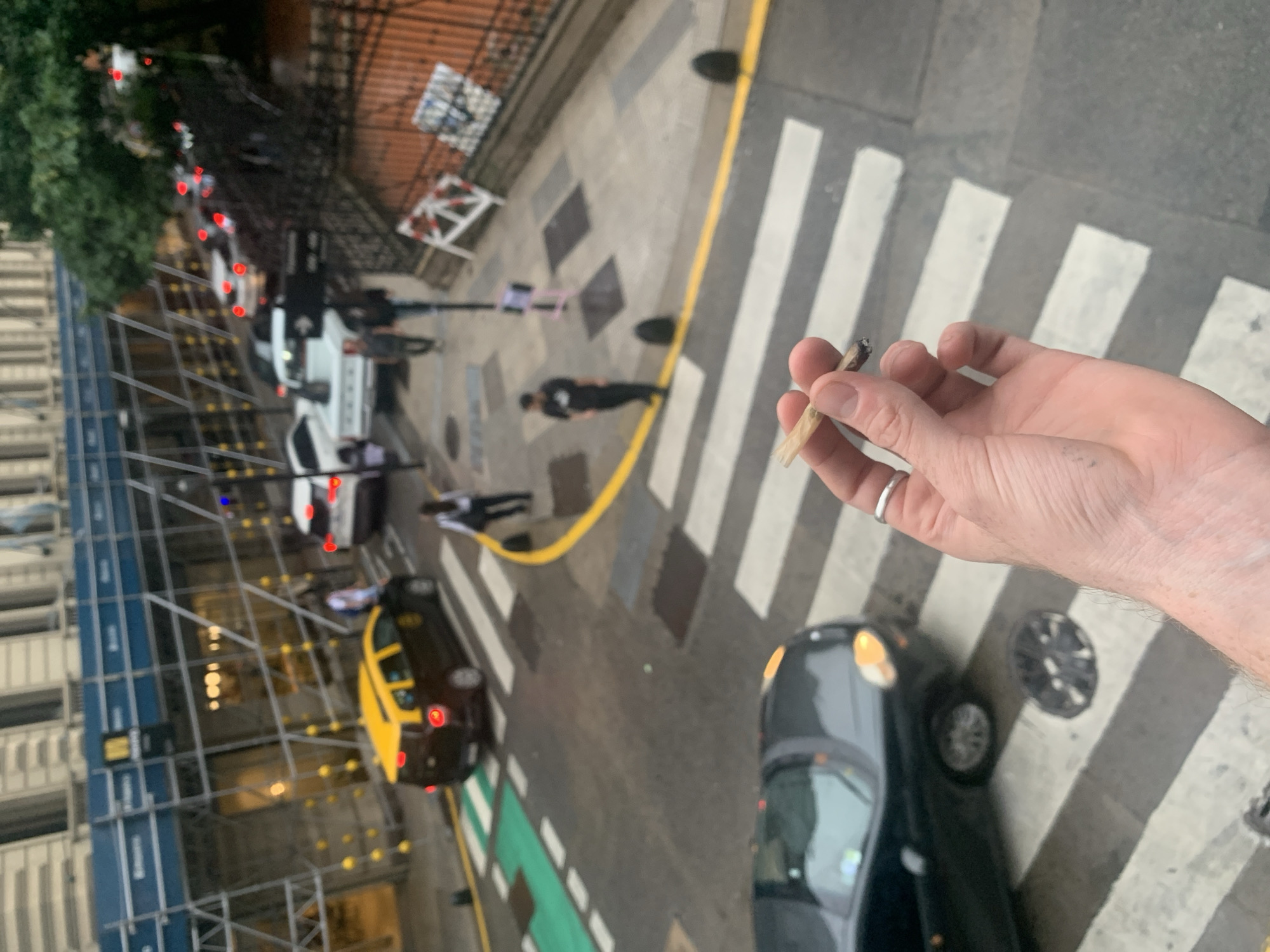 The image is an eye-level, tilted, high-angle shot of a hand holding a lit cigarette or joint, with a street scene in the background. The hand belongs to a light-skinned person wearing a silver ring on the ring finger. The street scene below is of Buenos Aires, Argentina, based on the presence of a yellow and black taxi cab, pedestrian crossing and street furniture. The street is wet, suggesting recent rain. Several people are walking on the sidewalks and crossing the street. Several cars are parked along the street. Some buildings on the left are under construction, with scaffolding. A yellow line is painted along the edge of the street, separating the road from the sidewalk. The overall image appears to be taken from a high floor of a building overlooking the street.
