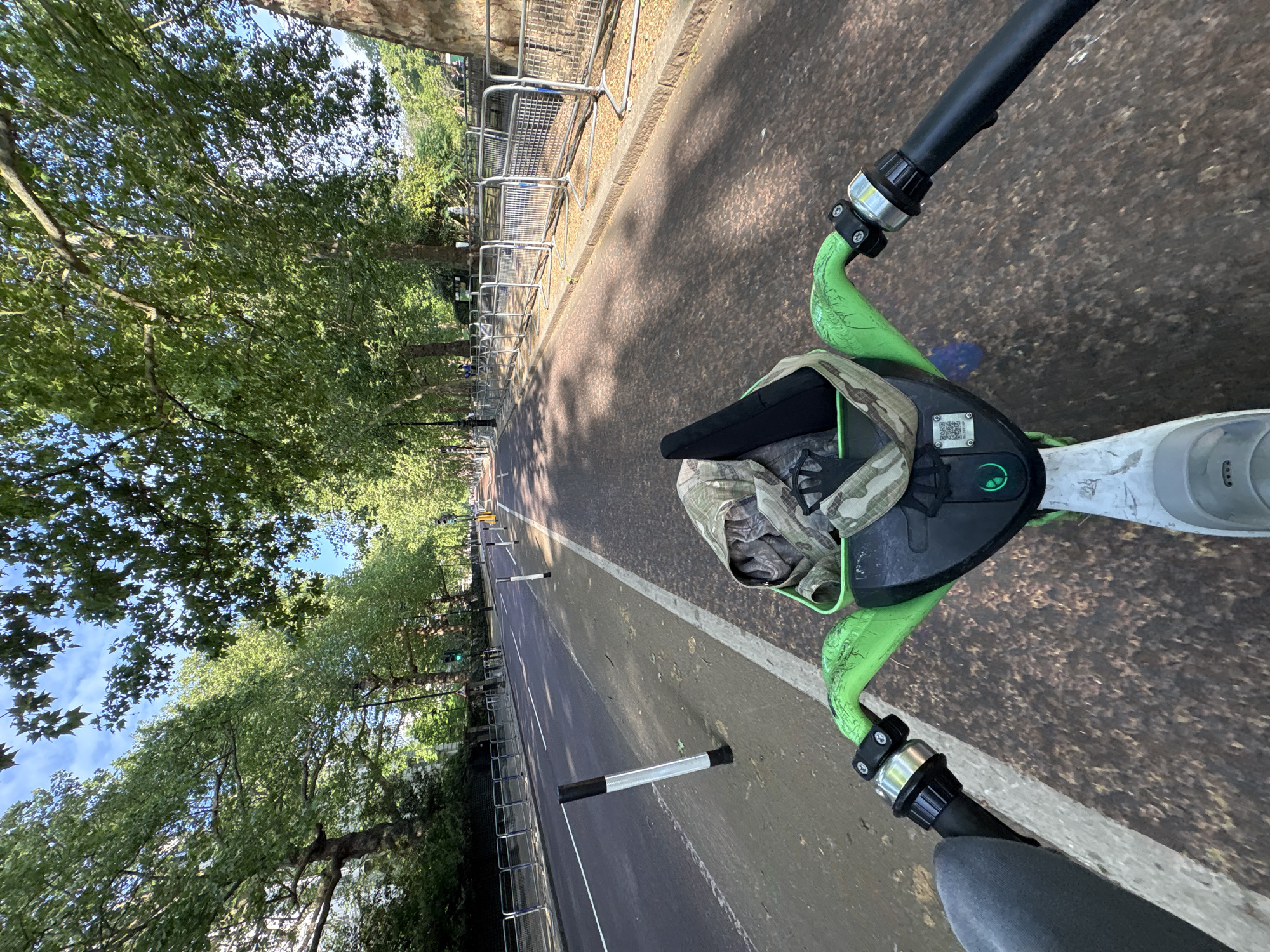 View from the handlebars of a green and black electric scooter, proceeding along a paved road in London, United Kingdom. The road is flanked by trees on the left and metal barriers on the right, with additional barriers further down the road. A camouflage-patterned soft item is partially visible on the front of the scooter. The road has a white line marking. Silver and black vertical bollards appear along the road. The sky is visible through the trees.