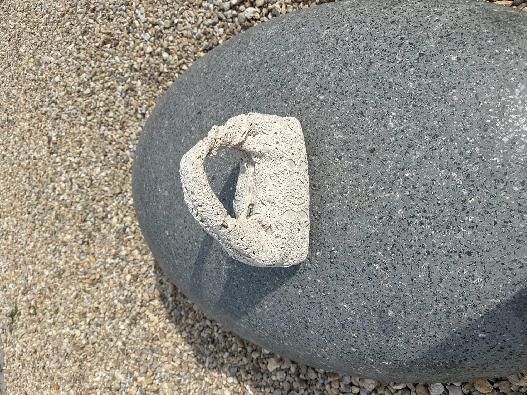 A small, cream-colored, crocheted handbag with a zipper closure rests on a large, smooth, gray, oval-shaped stone. The stone is positioned on a surface covered with small, light-colored pebbles. The lighting suggests it is daytime, with shadows cast on the stone. The background consists of more of the same pebbled surface. The location is the city of Yuzhnouralsk, Russia.
