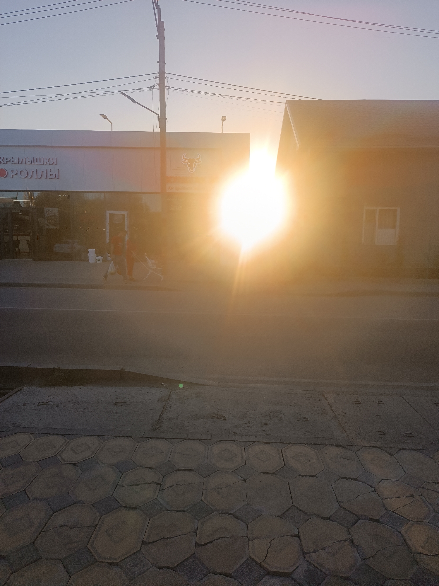 The image shows a street scene in Novopavlovka, Kyrgyzstan, at sunset. The sun is low in the sky, creating a bright glare that obscures details of the buildings and the road. In the foreground, a sidewalk with hexagonal paving stones is visible, with some cracks and wear. Beyond the sidewalk is a paved road. On the left side of the image, a building with a white facade has signage that appears to be in Cyrillic script. A bull logo is visible on the building. Further to the right, a darker building with a pitched roof and a window is present. Two people, appearing to be an adult and a child, are walking on the sidewalk near the building on the left, one pushing a stroller. A lamppost and power lines are visible in the sky.