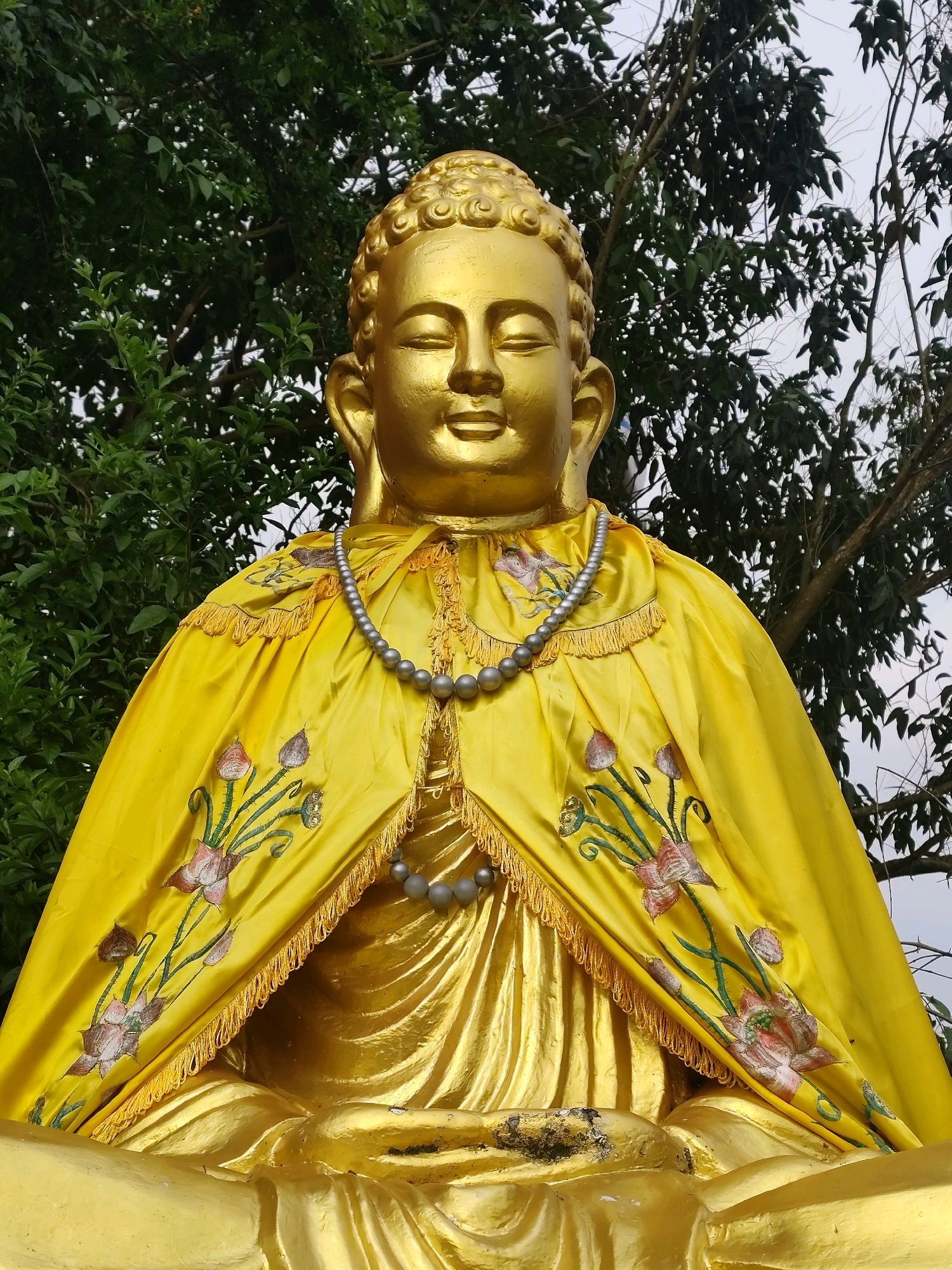A golden Buddha statue is prominently displayed in a seated meditation pose. The statue is adorned with a bright yellow robe that features embroidered floral patterns of pink and green lotuses and buds. A string of gray, bead-like ornaments is draped around the neck of the statue, with another strand visible lower down. The background consists of lush green foliage and tree branches against a bright, overcast sky. This scene is located at the Quan Âm Pagoda, situated on a hilltop overlooking the city of Lâm Viên - Đà Lạt, Vietnam, providing a serene and picturesque setting.