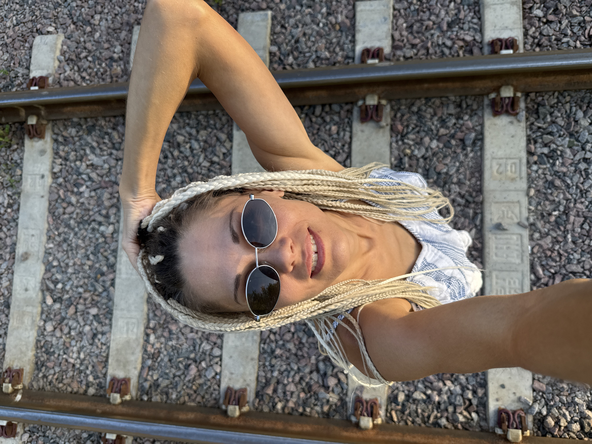 An overhead medium shot captures a woman with blonde braids posing on railroad tracks in Zhukovo, Russia. The woman, wearing sunglasses and a blue and white striped top, smiles at the camera with her left arm raised, touching her head. The railroad tracks are made of gravel and concrete sleepers. The lighting suggests it is daytime.