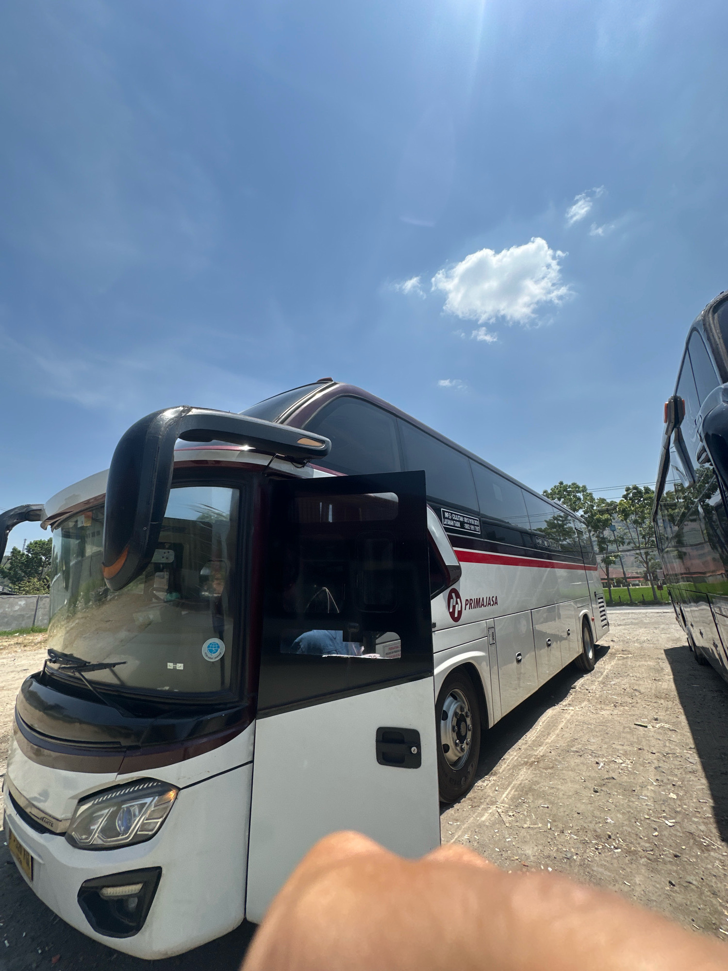 The image captures a bright, sunny day with a clear blue sky and a few wispy clouds. Two large white and maroon passenger buses are parked on a dusty, gravel lot, likely a depot or rest stop. The bus in the foreground is a PIMAJASA bus, identifiable by the prominent logo and text on its side. Its front passenger door is open, and a person is visible inside, possibly a driver or passenger. The bus features large windows, a prominent side mirror, and headlights. The second bus, parked to the right, is black and partially visible. The background shows a line of trees and some low-lying structures, suggesting an outdoor location. The overall scene is an unposed snapshot of buses in a lot, under a clear sky. The presence of the PIMAJASA branding and the context of the buses strongly suggests this image was taken at a PIMAJASA terminal or depot in or near Garut, Indonesia, a known hub for this bus company.