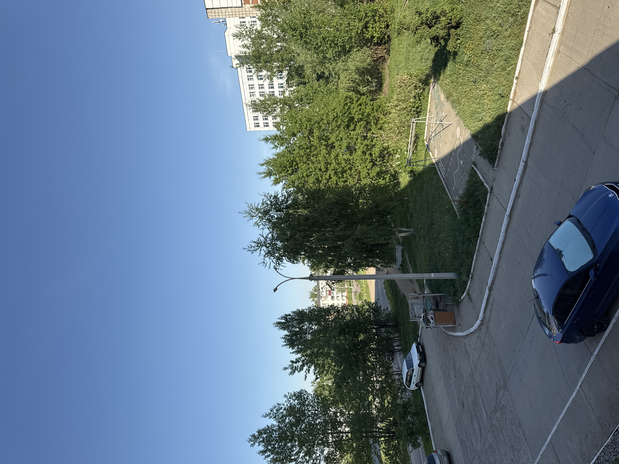 A slightly rotated high-angle view captures a residential street scene in Chaikovsky, Russia. A cloudless, bright blue sky dominates much of the left portion of the frame. On the right, a section of a concrete walkway runs along a grassy area; this area is marked by a worn, possibly child-painted playing surface. A dark blue sedan occupies the edge of the walkway, near a street. Further down the street are other parked vehicles, interspersed among the trees. A street light stands tall. In the background, building are visible, one white and high-rise. The other is smaller, white and red.
