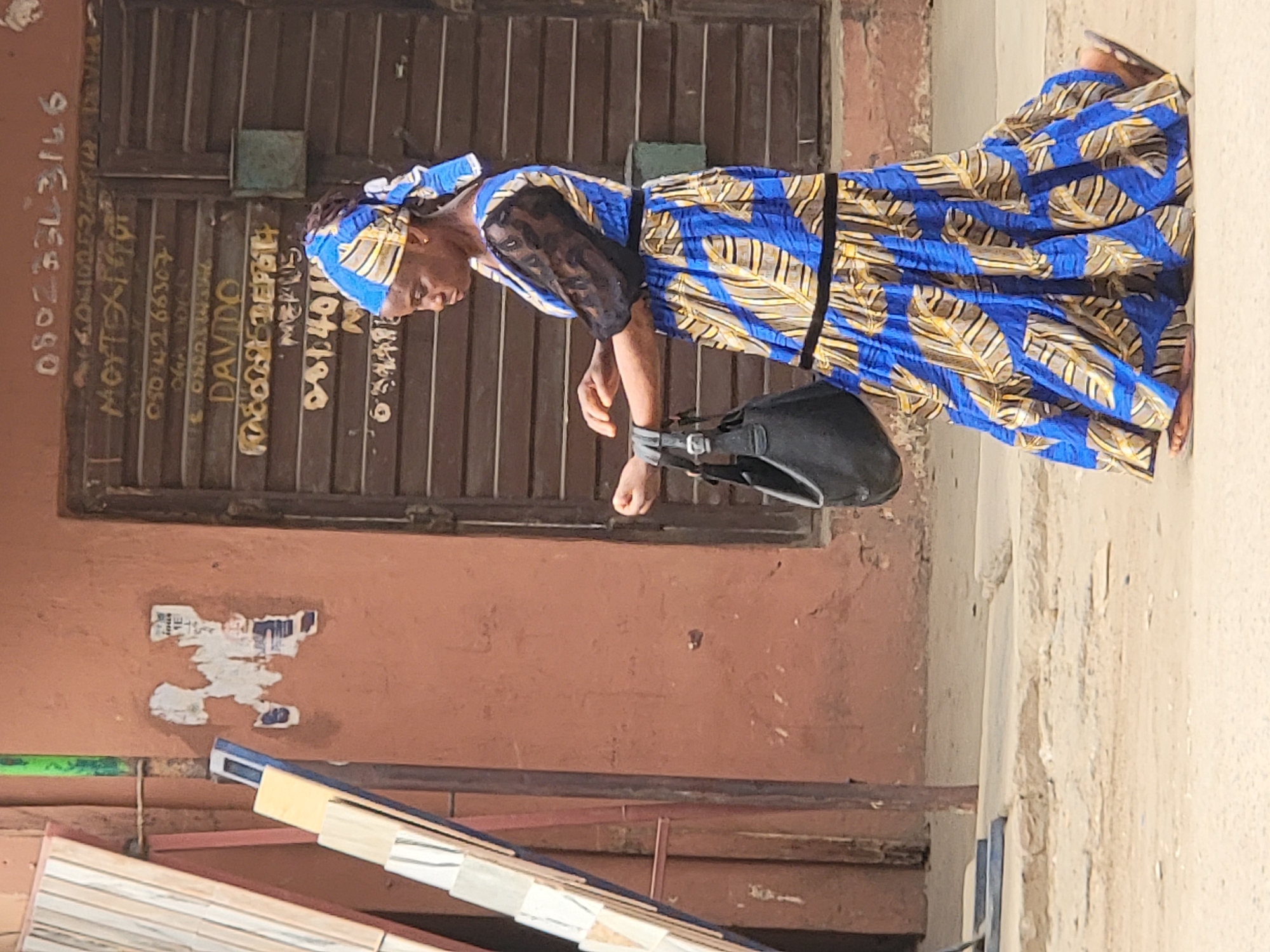 A woman wearing a vibrant blue, gold, and brown patterned traditional African dress and matching headwrap is captured in an outdoor setting in Maiburiji, Nigeria. She has a black, long-sleeved undershirt and a black belt cinching her waist. Her head is tilted downwards, and she appears to be looking at something or deep in thought, holding a black handbag with her right hand.
Behind her is a dark brown, slatted or corrugated metal door or gate covered with various handwritten white and yellow text, including phone numbers and names. Visible text includes "0802331316", "MOTEXT...", "08042 66307", "DAVIDO", "MEMUS", and "08140...". To the left is a reddish-brown wall with remnants of peeled posters or paint. In the lower foreground, construction materials or tiles are stacked on a stand. The ground on the right is a light-colored, possibly concrete or dirt surface. The bright lighting indicates it is daytime.