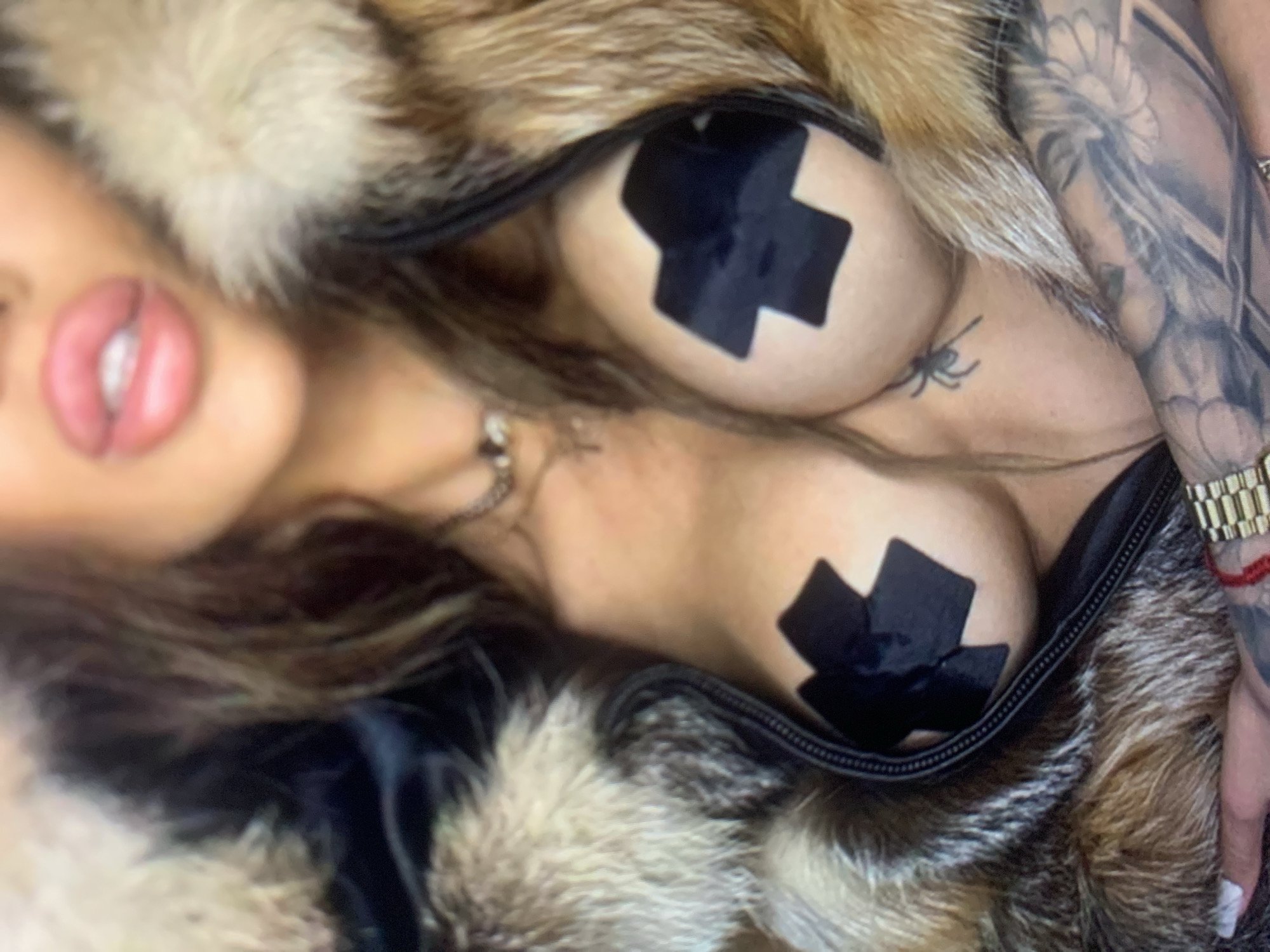 A close-up, medium shot shows a person from the chest up, wearing a fur-lined jacket. The person's breasts are partially visible and covered with black tape in a cross shape. They have tattoos on their chest and arms. The background is blurred, with hints of hair and the person's face. The lighting is soft and warm. The image is taken from a low angle.