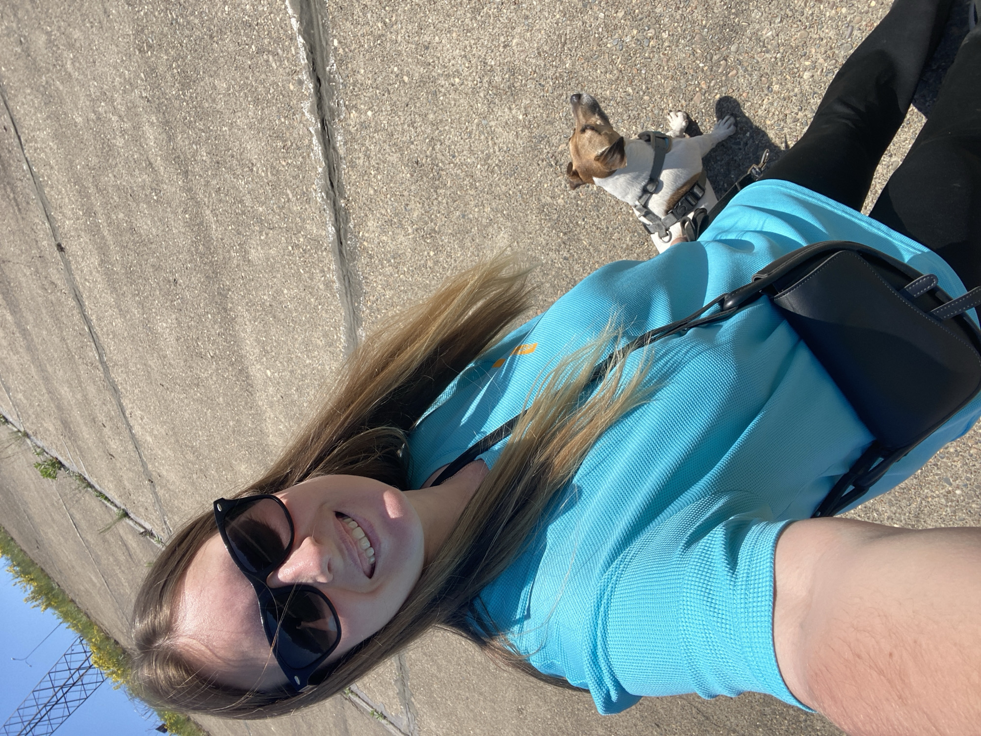 A woman is taking a selfie with her dog on a concrete surface, possibly a sidewalk. The woman is wearing a light blue shirt, black leggings, and sunglasses. She has a black purse slung over her shoulder. Her dog, a Jack Russell Terrier, is wearing a harness. The background includes a sidewalk with a crack and some greenery on the edge. In the distance, there are utility poles and a blue sky. The location is Noviy, Russia.
