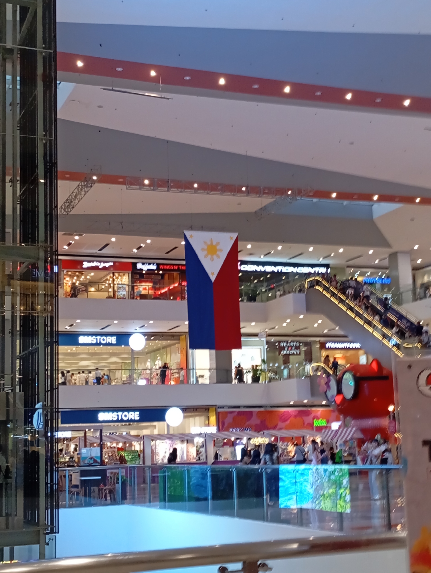 The image displays the interior of a brightly lit, multi-level shopping mall, likely in Davao City, Philippines, as indicated by the prominent Philippine flag hanging vertically from the ceiling in the central mid-ground.
Numerous people are visible throughout the mall, engaging in various activities such as walking, shopping, and riding escalators. An escalator on the right side of the image is packed with individuals ascending to an upper floor. The mall features modern architecture with sleek railings and large open spaces.
Several stores and establishments are identifiable by their signage. On an upper floor, a large sign for "CONVENTION CENTER" is visible. Below this, a "SM STORE" sign appears prominently, with another "SM STORE" sign on a lower level. Other visible store names include "Burger & Jamm," "Buffalord Wings N' Th," "HEARTS & ARROWS," and "STRAIGHTUP (Davao)." On the lowest visible level, there are food stalls or cafes with striped awnings, with partial names like "Koloko" and "café e" readable. A large, red, cartoon-like figure with big eyes stands on the lower right, possibly a mascot or part of an attraction.
The overall scene depicts a bustling commercial environment. The lighting is uniform and bright, typical of an indoor mall during its operating hours, making the exact time of day difficult to determine. A glass-paneled structure, possibly an elevator, runs vertically along the left side of the frame.