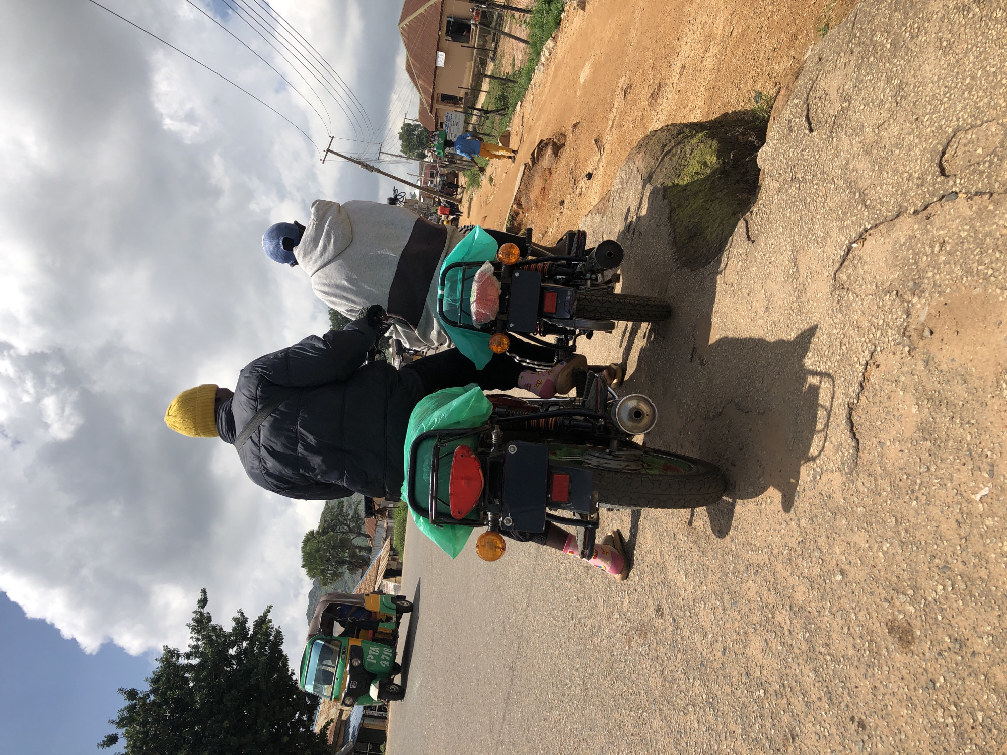 The image depicts a street scene in Jos, Nigeria, characterized by a dusty, uneven asphalt road with a dirt shoulder. In the foreground, two people are riding on a motorcycle, viewed from behind. The rider wears a dark jacket and a yellow beanie, while the passenger sports a grey hoodie and a blue cap. Both are positioned on the motorcycle, which has a green tarp covering its rear. The motorcycle is in motion, as indicated by the shadow it casts.
Further down the road, a three-wheeled auto-rickshaw, painted green and yellow, is visible. It has the inscription "PTA 4718" on its side and appears to be carrying passengers. In the background, a line of buildings with corrugated iron roofs suggests a residential or commercial area. Telephone poles and wires are present, typical of an urban environment. The sky is partly cloudy, with large white cumulus clouds against a bright blue backdrop. The overall impression is a snapshot of daily life and transportation in a Nigerian city.