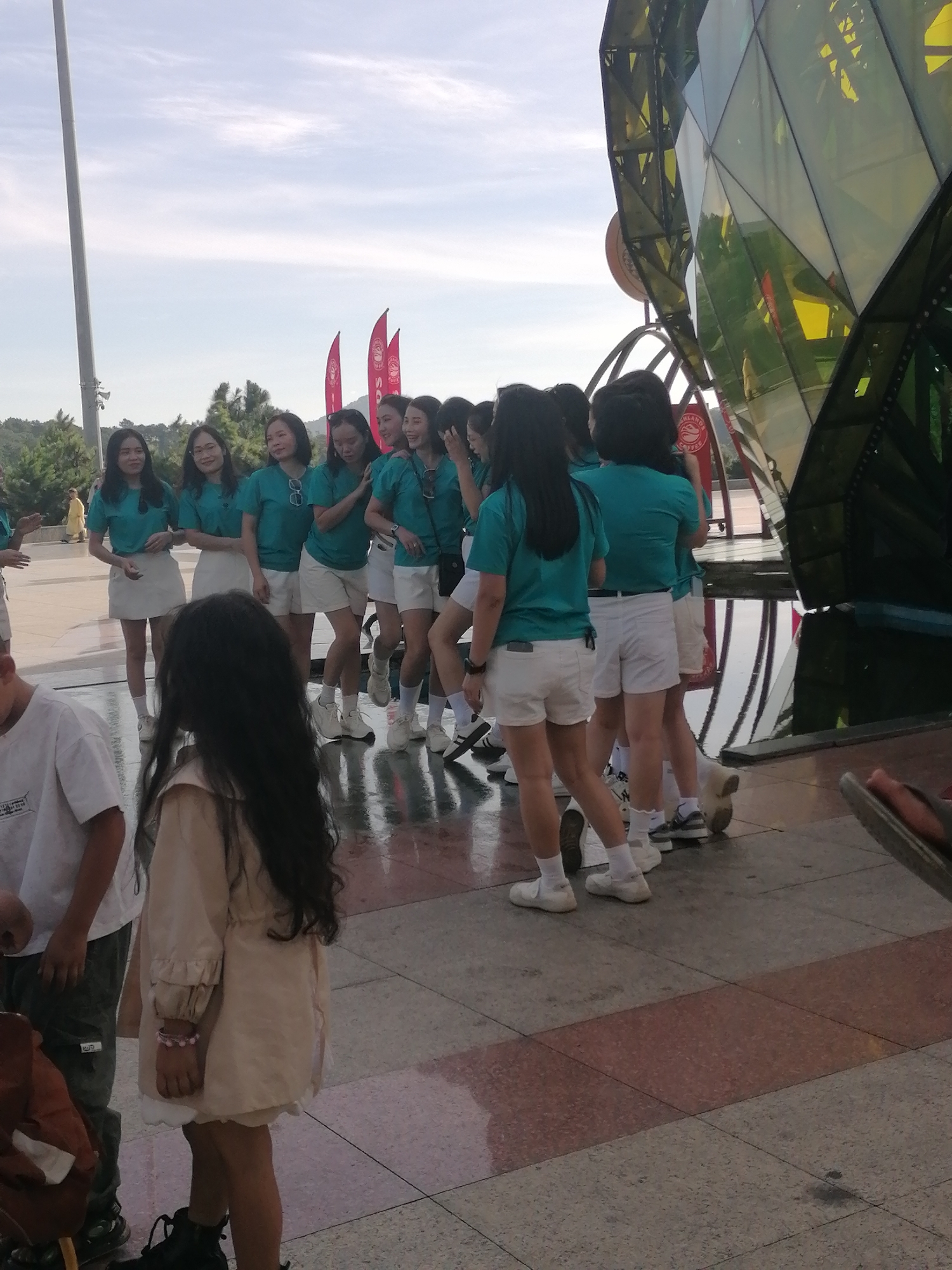 A group of young women are posing for a photo in front of a large, geometric glass structure. They are all wearing matching teal t-shirts and white shorts or skirts. Some are smiling and looking at the camera, while others are looking in different directions. The women are standing on a paved area with a red and pink patterned section. In the background, there are trees, a tall pole with lights, and a bright sky. The glass structure appears to be a modern building with angular panels. The scene suggests a casual outdoor gathering or event. The location is likely the Da Lat Flower Park, a popular tourist attraction in Da Lat, Vietnam, known for its floral displays and modern architectural elements, including the iconic Geometric Dome structure which this glass building resembles.