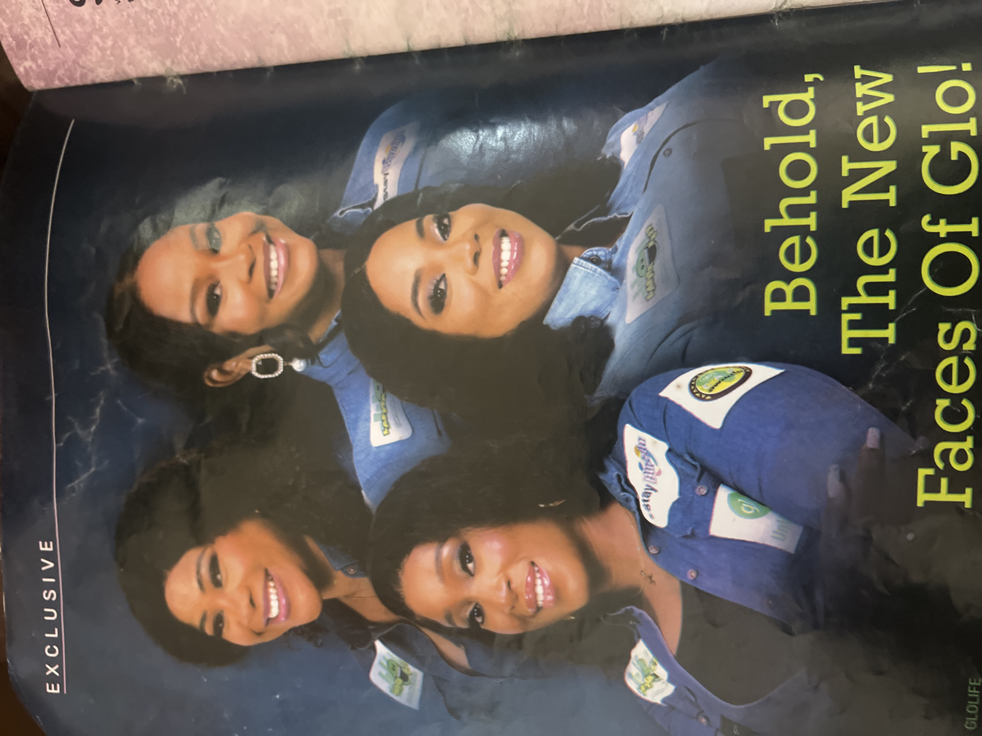 The image depicts a magazine cover with the title "Behold, The New Faces Of Glo!". Four women are shown, all appearing to be of Nigerian descent, with dark hair and smiling. They are all wearing matching denim shirts adorned with various patches. The woman on the far left has a patch on her shirt that reads "Stay Bling". Another patch on the same shirt features a cartoon character. The woman in the center-left also has a "Stay Bling" patch. The woman in the center-right has a patch with a green circle and the word "Glo" inside. The woman on the far right has a patch that appears to have a green circle and possibly a cartoon character. She is also wearing a prominent silver earring with rhinestones. The background of the magazine cover is a dark blue, possibly representing a night sky or a studio backdrop. The text "GLOLIFE" is visible in the upper left corner, and "EXCLUSIVE" is written vertically on the right side. There is no specific indication of the location being Jalingo, Nigeria, or any other city.