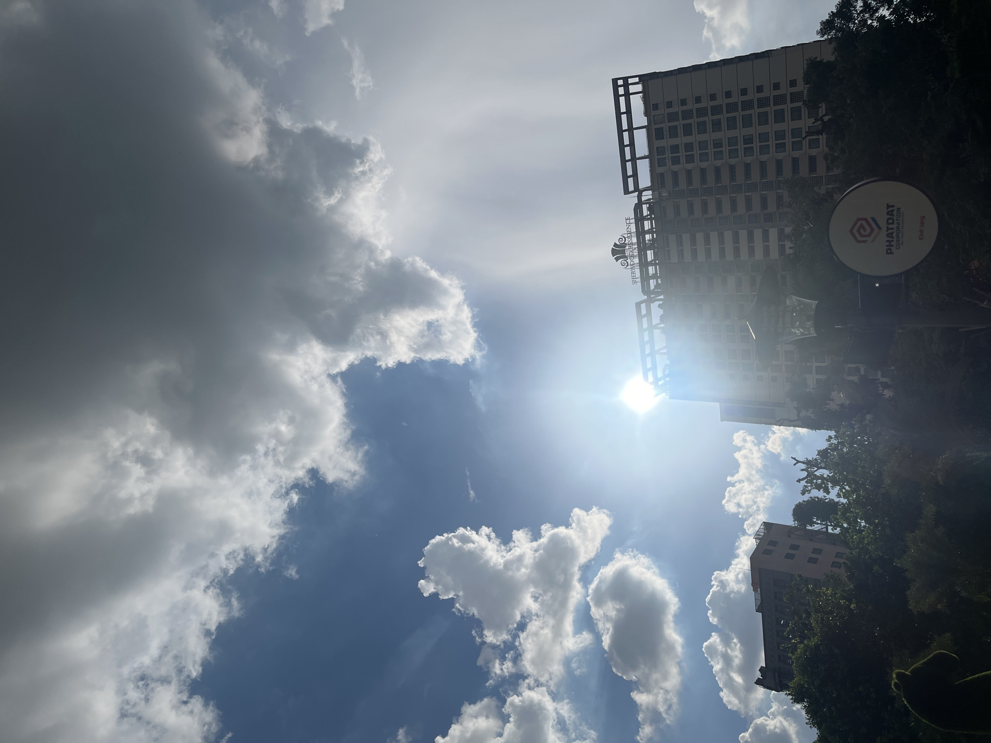 The image captures a view of the Sherwood Residence building in Thủ Đức City, Vietnam, under a dramatic sky. The sun shines brightly, its rays partially obscured by thick, cumulus clouds. To the left, lighter white clouds contrast with darker, heavier gray clouds dominating the upper right portion of the frame, suggesting an approaching storm or the aftermath of one. The architecture of the Sherwood Residence, a tall, modern building with multiple floors and a distinctive top structure, is visible. Green foliage and trees partially obscure the lower sections of the building and the surrounding area, indicating a densely vegetated urban environment. A streetlamp and a circular sign with the "PHATDAT CORPORATION" logo are also present in the foreground, adding context to the urban setting. The scene is captured from a low-angle perspective, emphasizing the scale of the building against the vast sky.