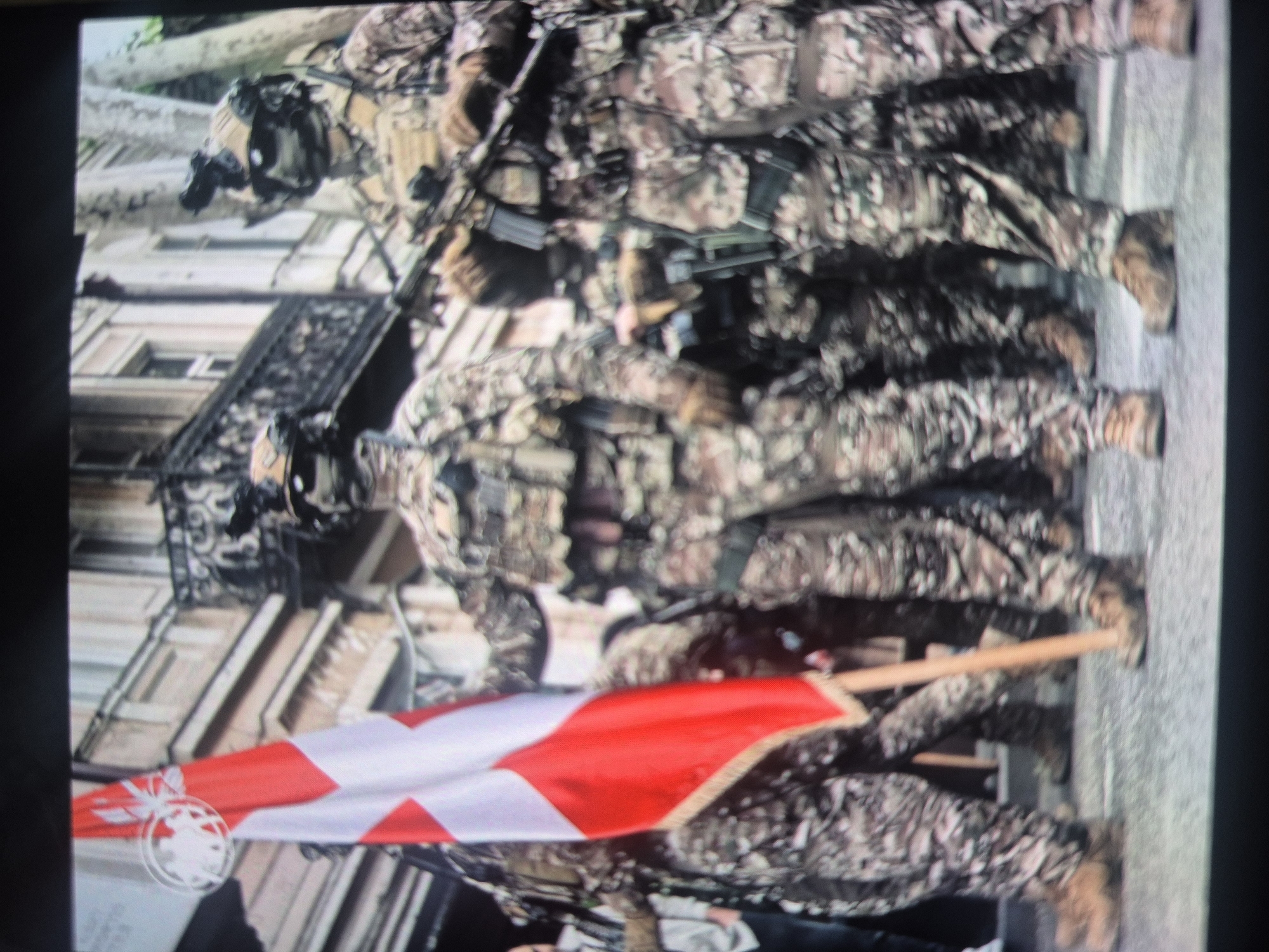 The image shows three soldiers in camouflage uniforms marching on a street in Tbilisi, Georgia. The soldiers are carrying rifles. One soldier is carrying a flag with a red cross on a white background. Buildings are visible in the background.
