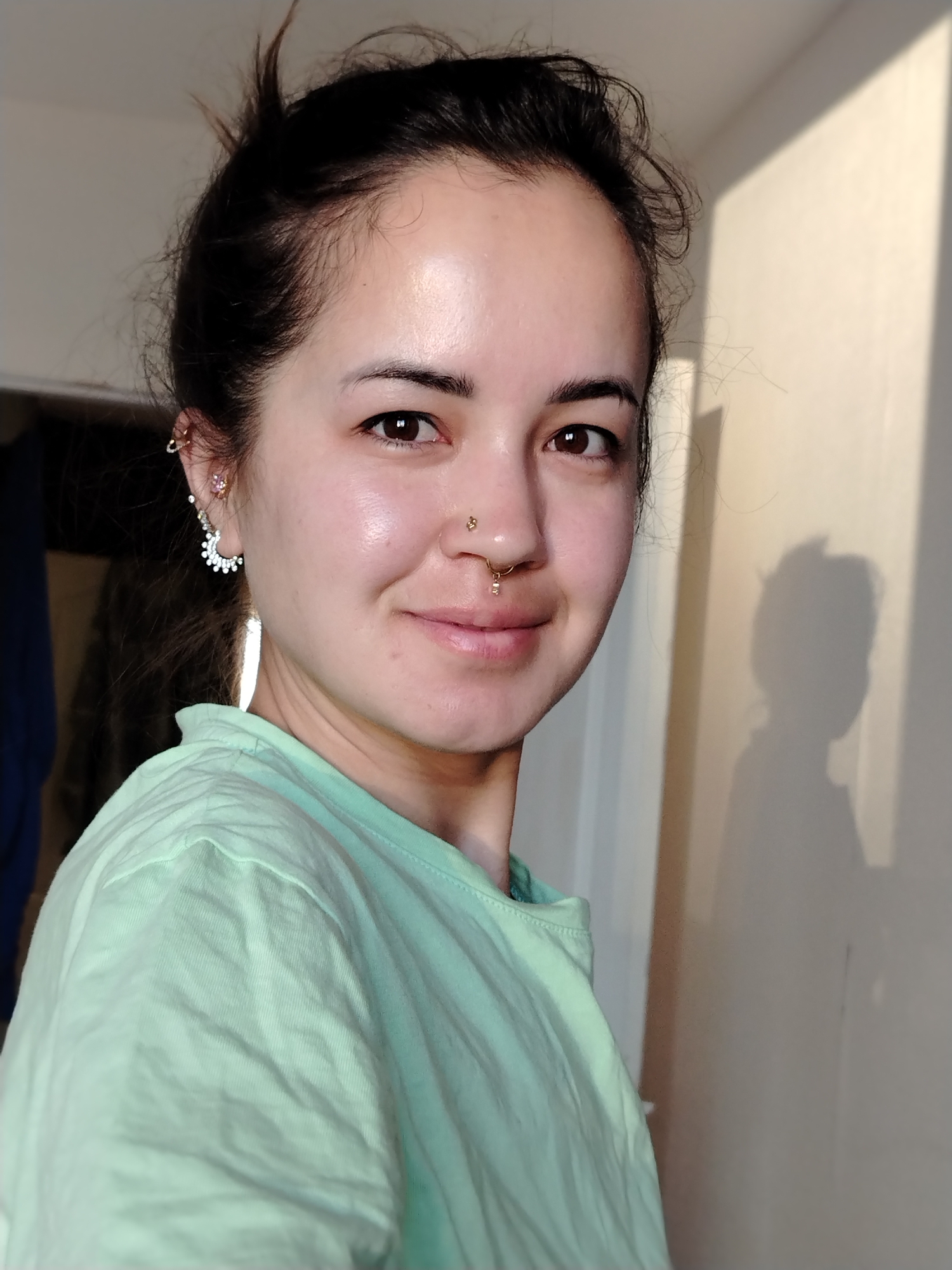 A medium-shot photograph depicts a woman with brown hair pulled up in a loose bun. She is fair-skinned and has brown eyes, adorned with two nose piercings. She wears a light green t-shirt. Her right ear is pierced with multiple earrings. To the right of the woman is a cream-colored wall with a shadow cast upon it. No information is available to confirm the location in the city of Noviy, Russia.