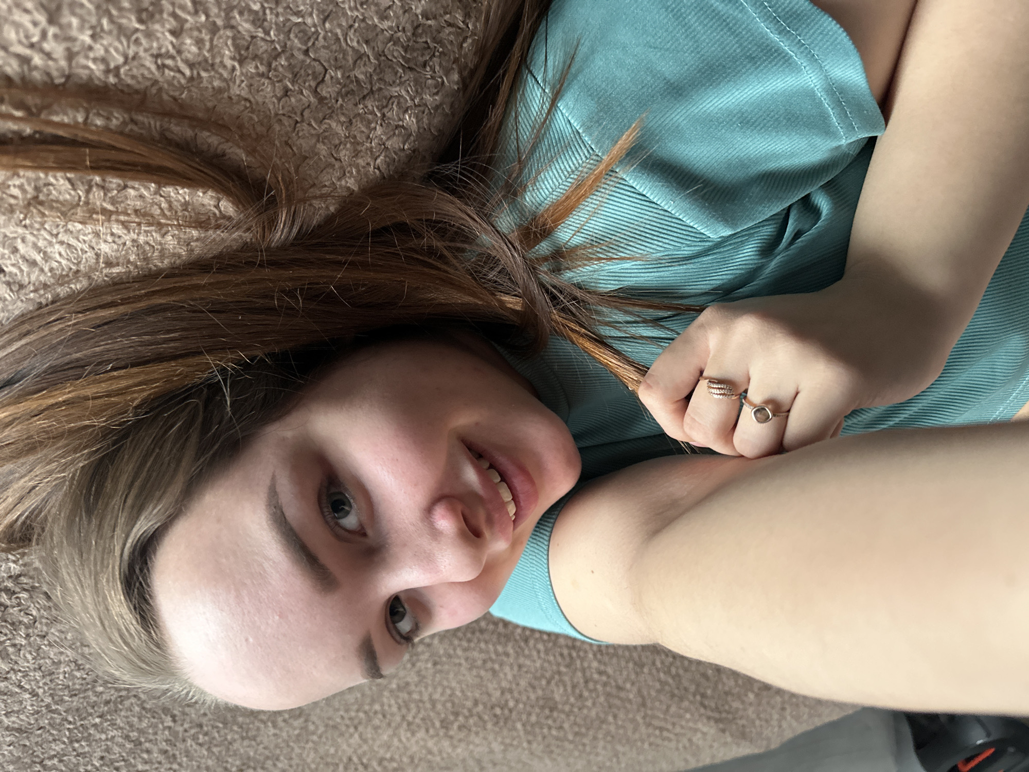 A woman is lying on a brown shag carpet. She is wearing a teal short-sleeved shirt and two rings on her left hand. Her hair is light brown with red highlights. The location is consistent with an indoor setting. The city of Chaikovsky, Russia, cannot be determined from the image.