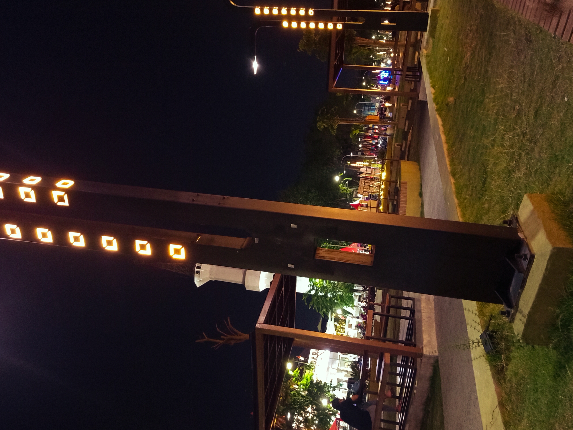 The image depicts an outdoor scene at night, likely in Sidoarjo, Indonesia, given the context. A large, dark, rectangular structure with illuminated diamond-shaped openings runs vertically down the right side of the frame. To the left, a covered seating area with wooden tables and benches is visible, with a person sitting at one of the tables. Beyond this, there are buildings with illuminated signage, including one that reads "SUNDAR." A white tower, possibly a minaret, is also visible in the background. The scene is lit by various artificial lights, including streetlights and decorative lights. The ground is a mix of pavement, grass, and gravel. The overall impression is that of a public space or a commercial area illuminated for nighttime activity.