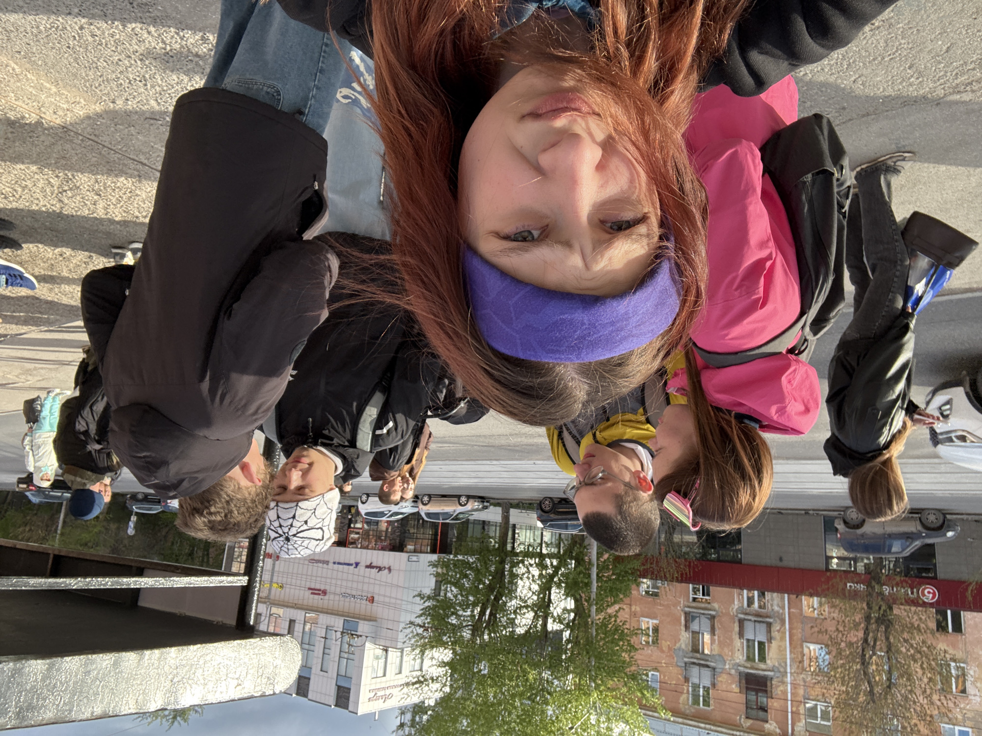A selfie taken in Perm, Russia, shows a group of people. The selfie taker, a woman with red hair and a purple headband, is in the foreground. Behind her are several other people, including a person wearing a white hat with a spiderweb pattern, another wearing glasses, and a person in a pink jacket. The background features city buildings and trees. A building with "Assays" written on it and a building with a large number "5" are visible. Several cars are parked on the street.