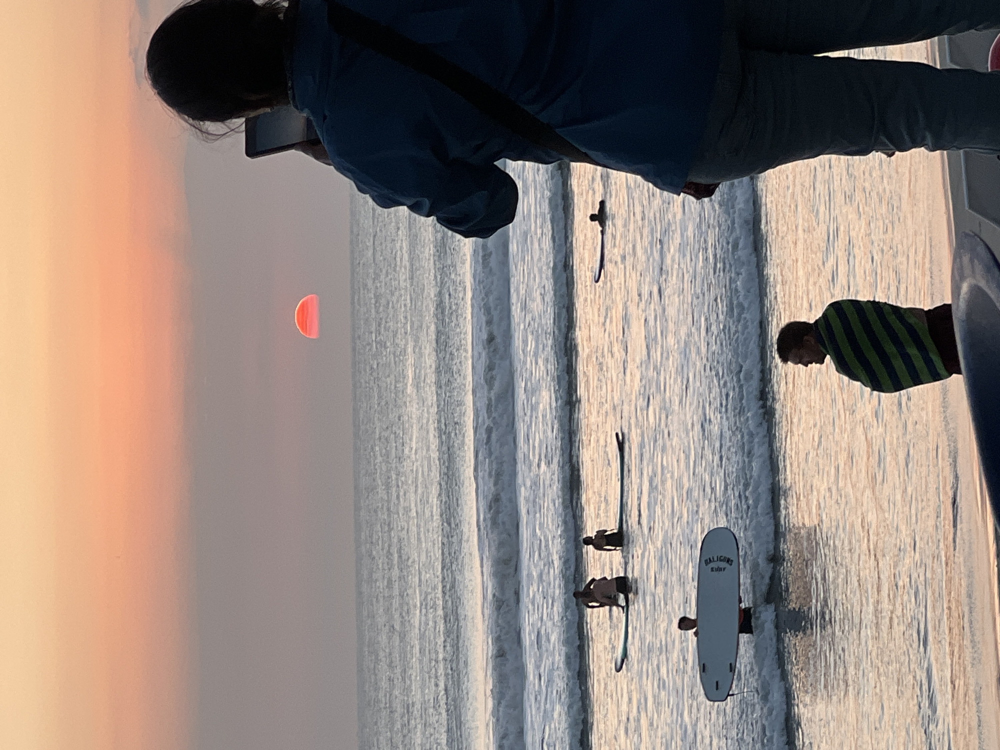 The image depicts a sunset over the ocean at Kuta Beach, Indonesia. In the foreground, a person in a blue jacket and grey pants stands facing the ocean, holding a tablet or phone to take a picture. Their back is to the viewer. Further out in the water, several individuals are on surfboards. One person stands holding a white surfboard. Two other figures are seated on surfboards. The sun is setting, casting an orange glow across the sky and water. On the beach, a man wrapped in a green and blue striped towel stands facing the ocean. The overall scene is one of a tranquil evening at the beach.