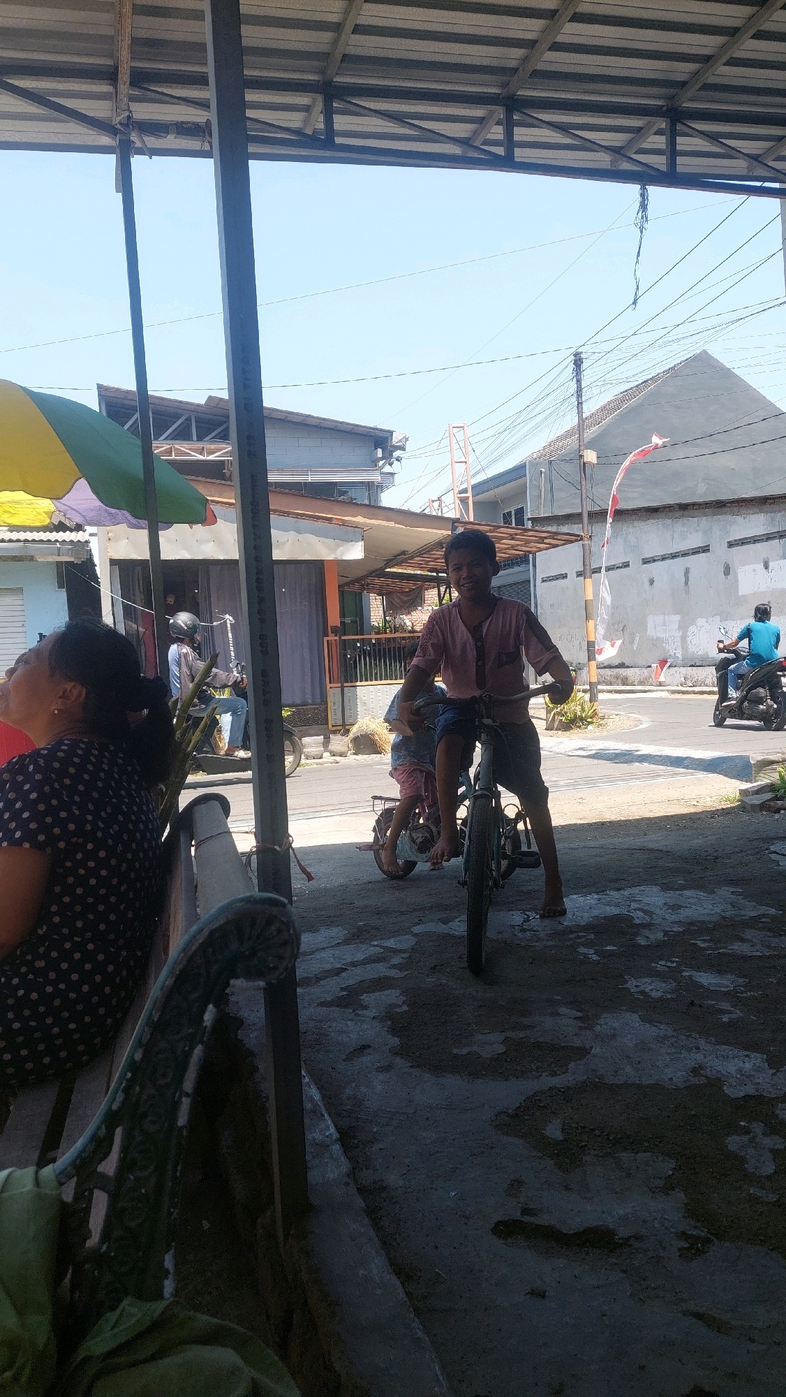 In Sidoarjo, Indonesia, under a corrugated metal roof supported by metal poles, a young boy in a pink shirt rides a bicycle with a smaller child sitting behind him. The boy grins at the camera, his bare feet on the pedals. In the background, a woman with dark hair wearing a polka-dot shirt sits on a dark bench to the left. To the right, a man in a blue shirt rides a motorcycle. Buildings with light-colored walls and tiled roofs line the street, with electrical wires crisscrossing overhead and a red and white flag flying from a pole. The ground is a mix of concrete and patches of dark, wet-looking material, suggesting recent rain. The scene is outdoors on a bright, sunny day.