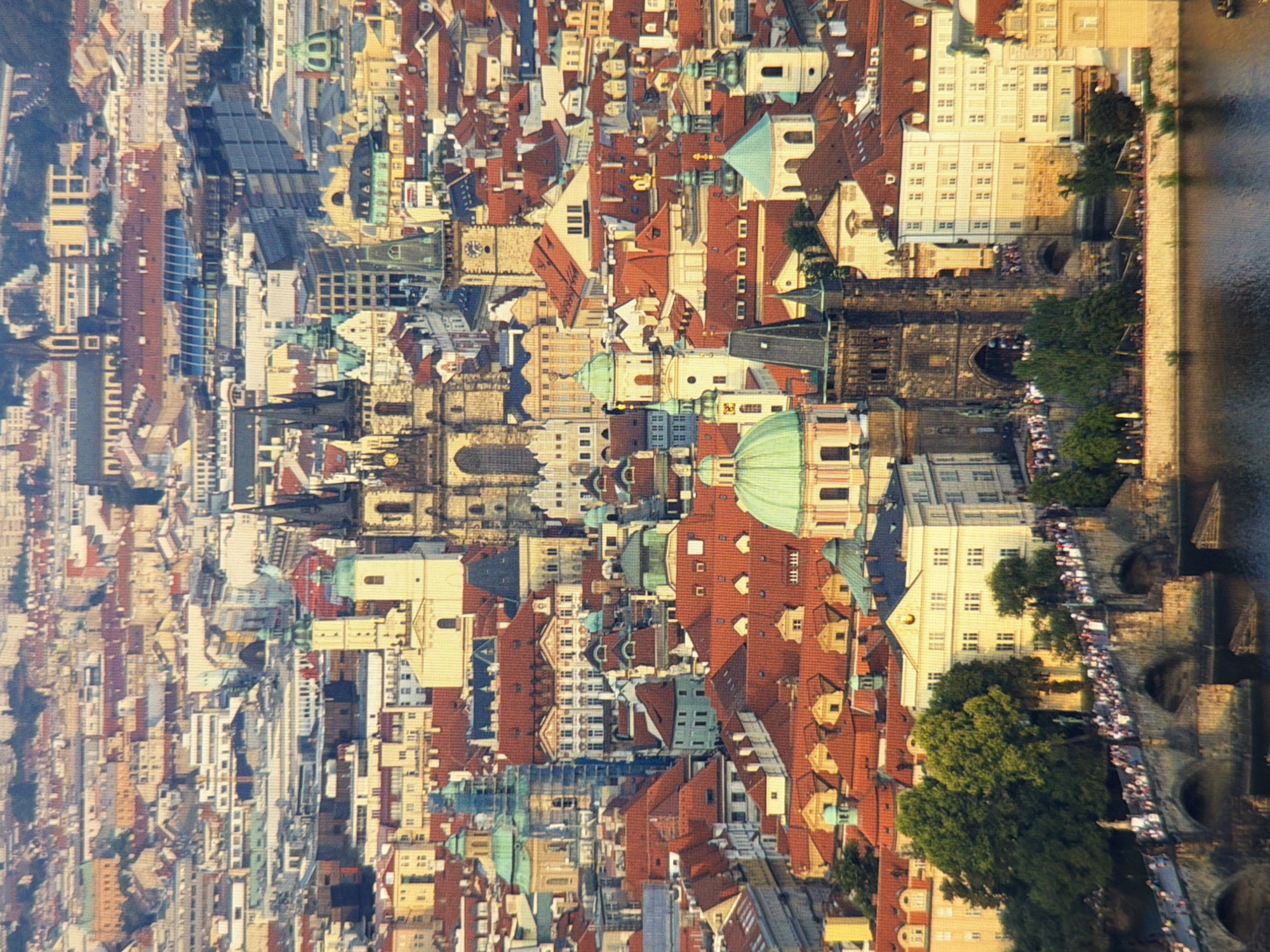 This is an aerial, sunlit photograph of a dense European cityscape. The architecture is predominantly red-tiled roofs and light-colored buildings with numerous spires and domed structures. Prominent in the foreground is a stone bridge with multiple arches, crowded with pedestrians. Beyond the bridge, more buildings rise, including a large, dark stone tower with an arched entryway. The river in the very bottom of the frame reflects the muted colors of the sky and surrounding structures. The city appears to be experiencing a warm day, possibly late afternoon, given the long shadows.
The image depicts Prague, Czech Republic, not Kyiv, Ukraine. Specifically, the iconic Charles Bridge with the Old Town Bridge Tower is visible, as well as the Church of Our Lady before Týn and other historic buildings in the Old Town Square.